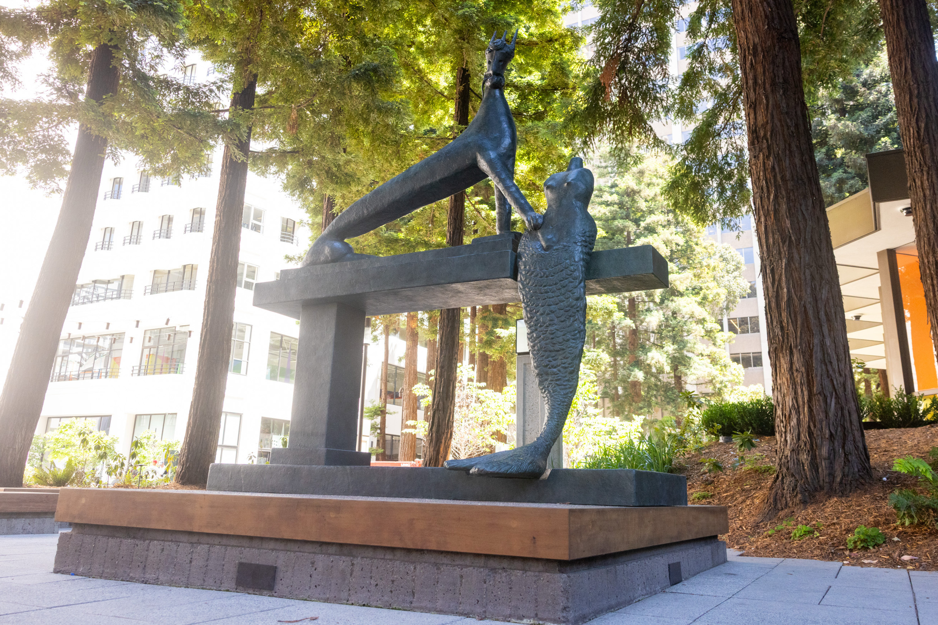 The image shows a modern sculpture of two mermaid-like figures meeting at a crossbeam, surrounded by tall trees and buildings in a sunny outdoor plaza.