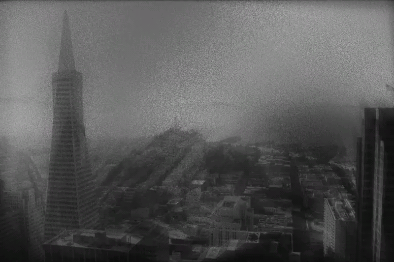 The image depicts a grainy, black-and-white view of a city skyline. A tall, pyramid-shaped building stands prominently on the left, with fog enveloping the background.