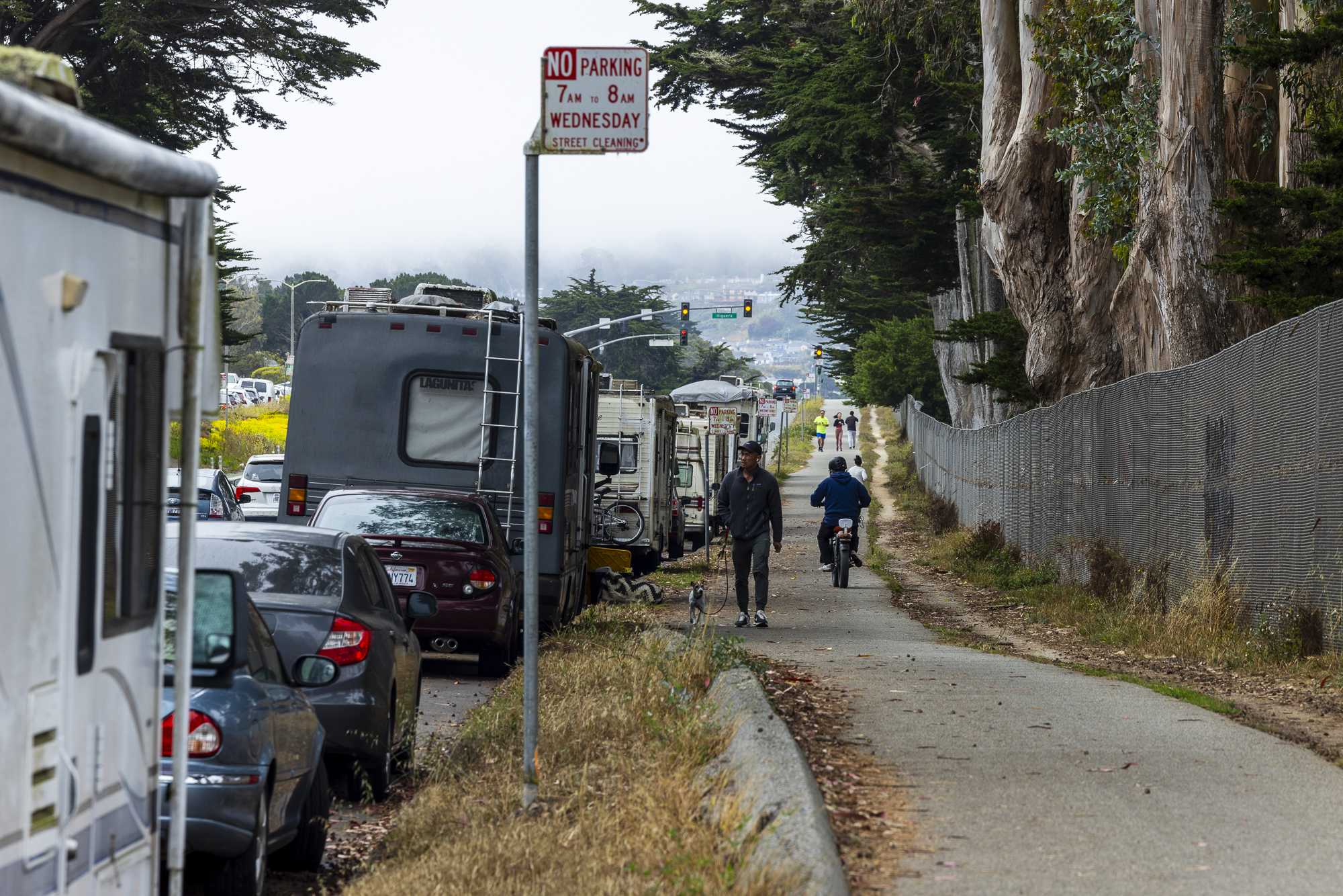 A narrow path runs beside parked cars and RVs on a street lined with tall trees. People walk and cycle along the path under a cloudy sky.