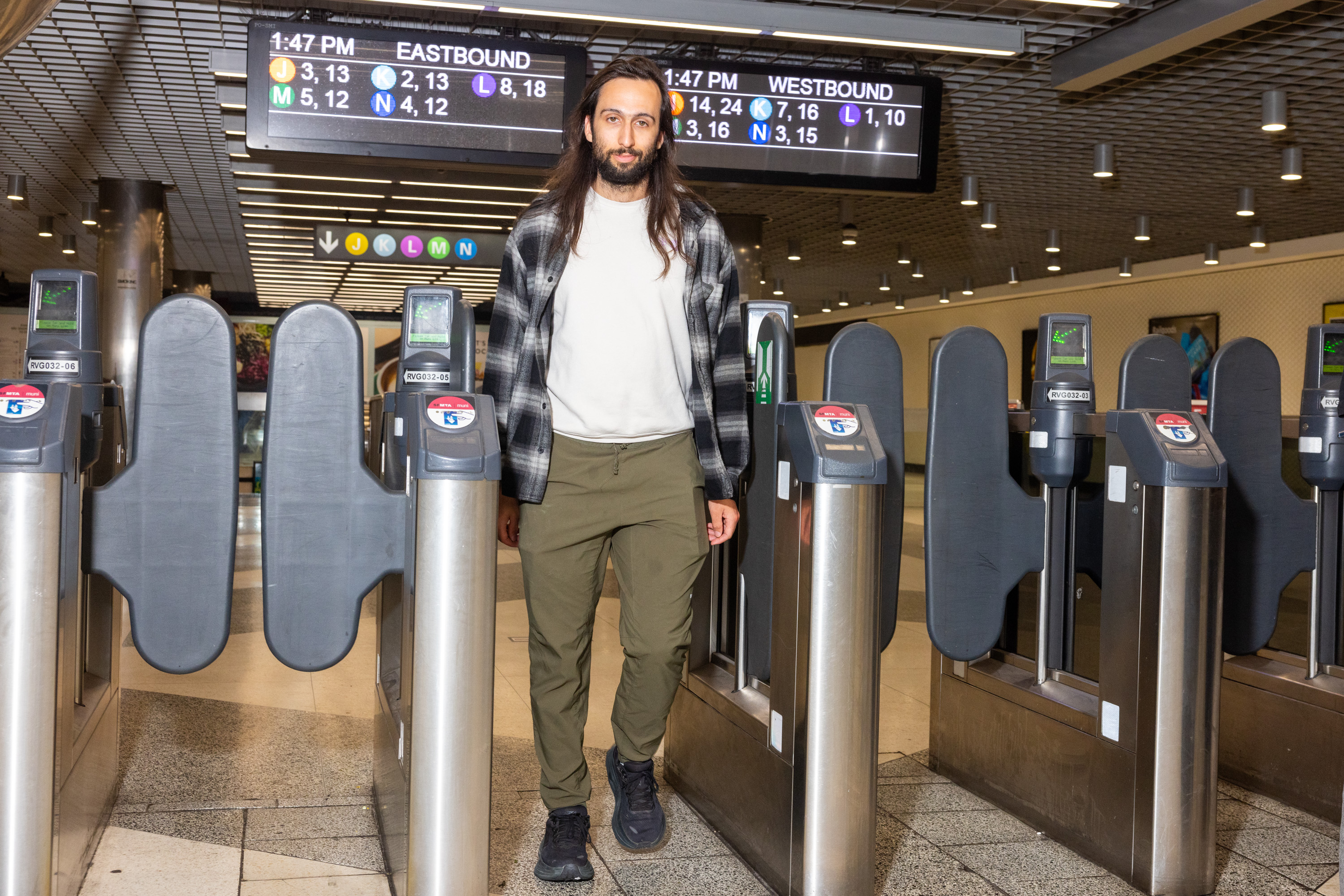 A person with long hair and a beard walks through a subway turnstile. They wear a plaid shirt over a white top and olive pants. There are subway signs above.