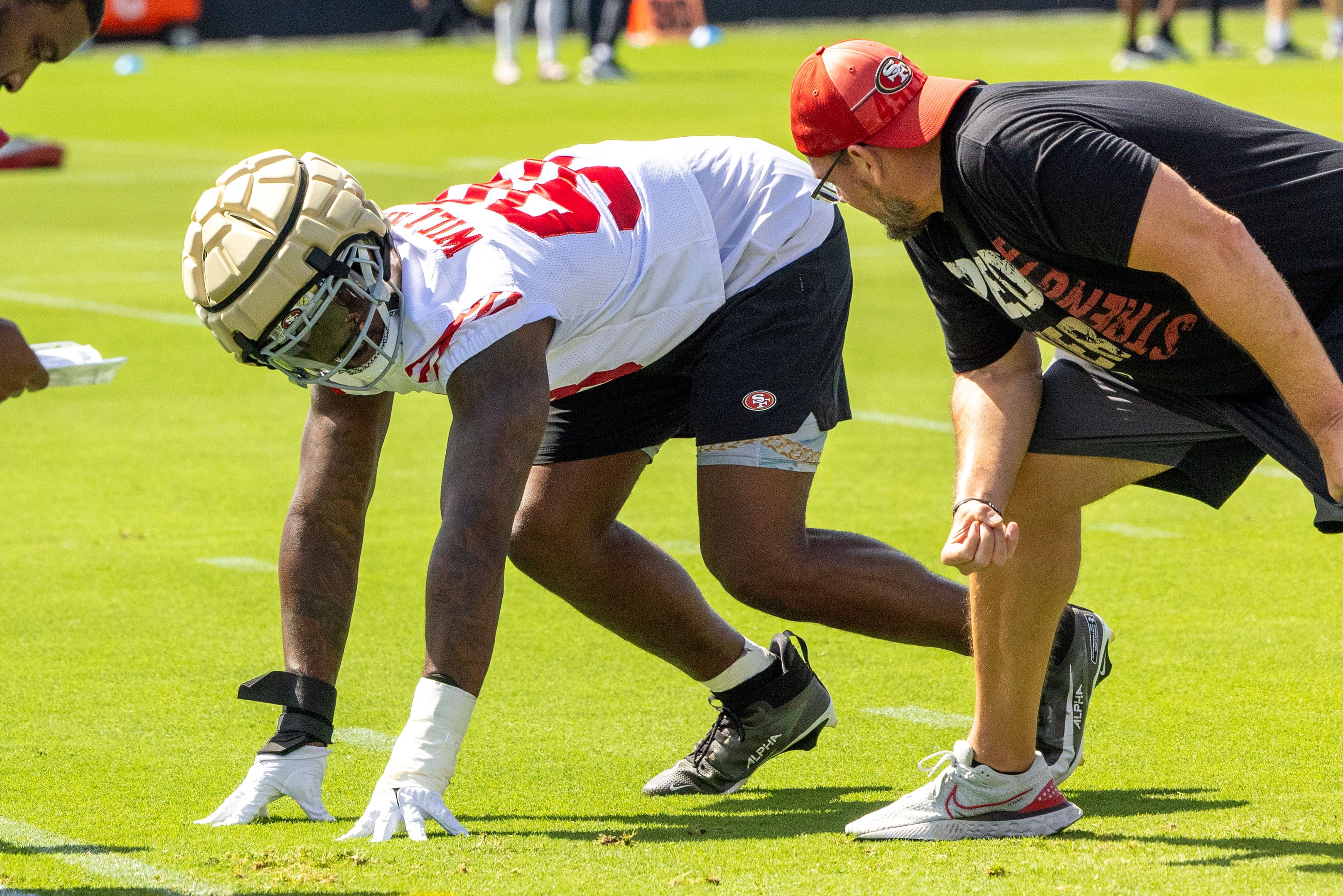 A football player in a white jersey and padded helmet crouches on a field, preparing to start. A coach in a red cap and black shirt kneels beside him.
