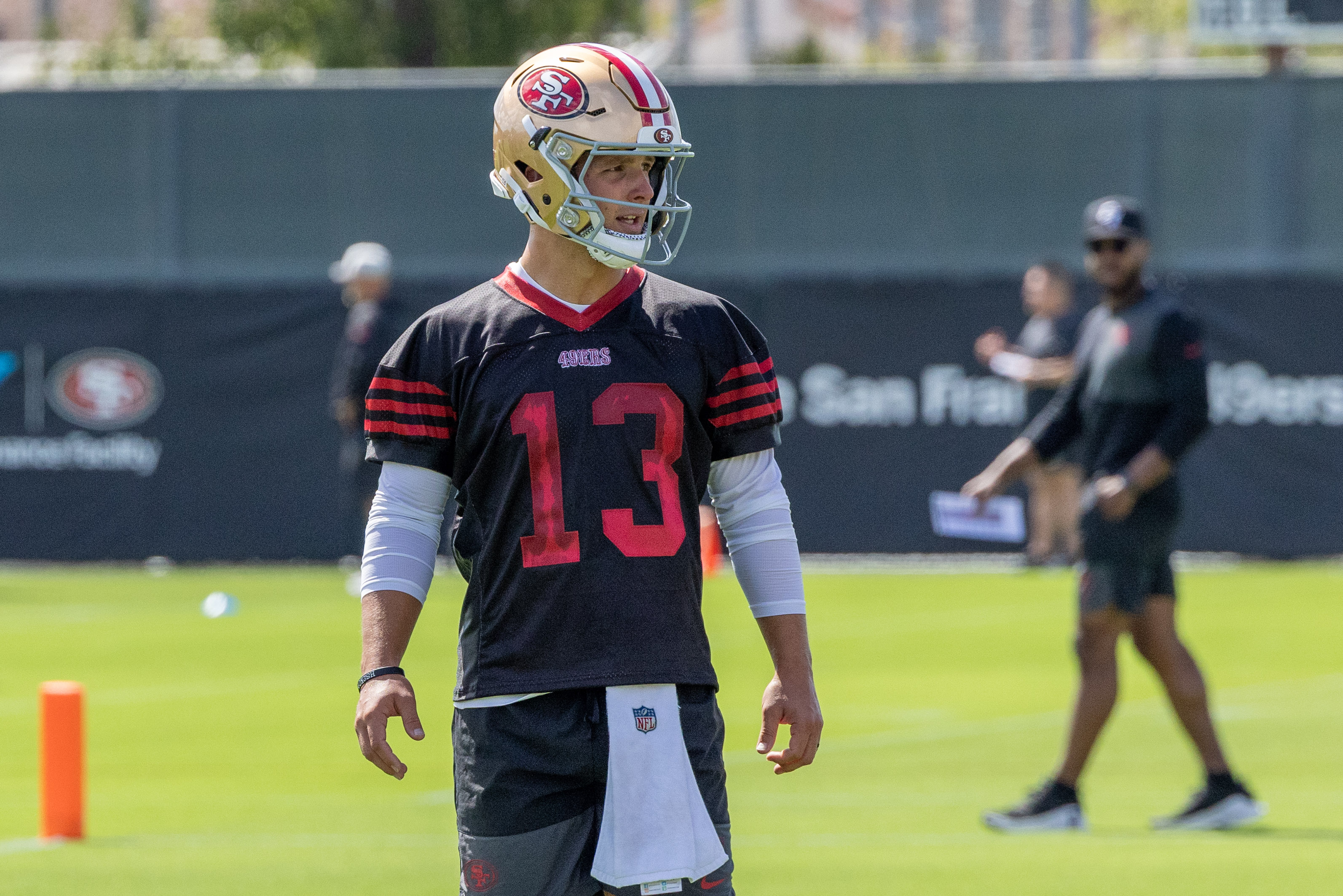 A football player in a 49ers uniform and helmet stands on a practice field, wearing jersey number 13. Others are in the background with a blurred banner.