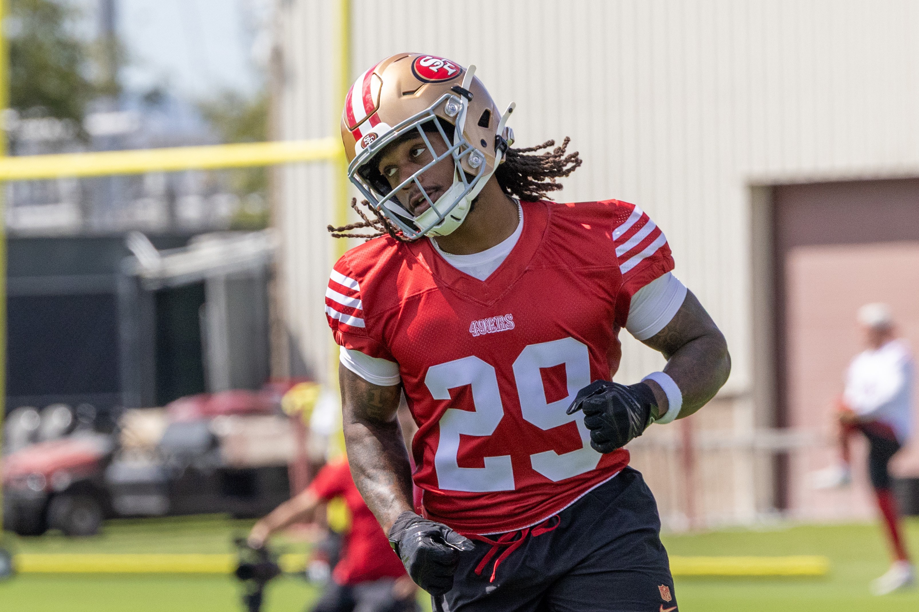 A football player wearing a red jersey with the number 29 and a gold helmet is on a field. They are mid-action, and there are blurred figures in the background.