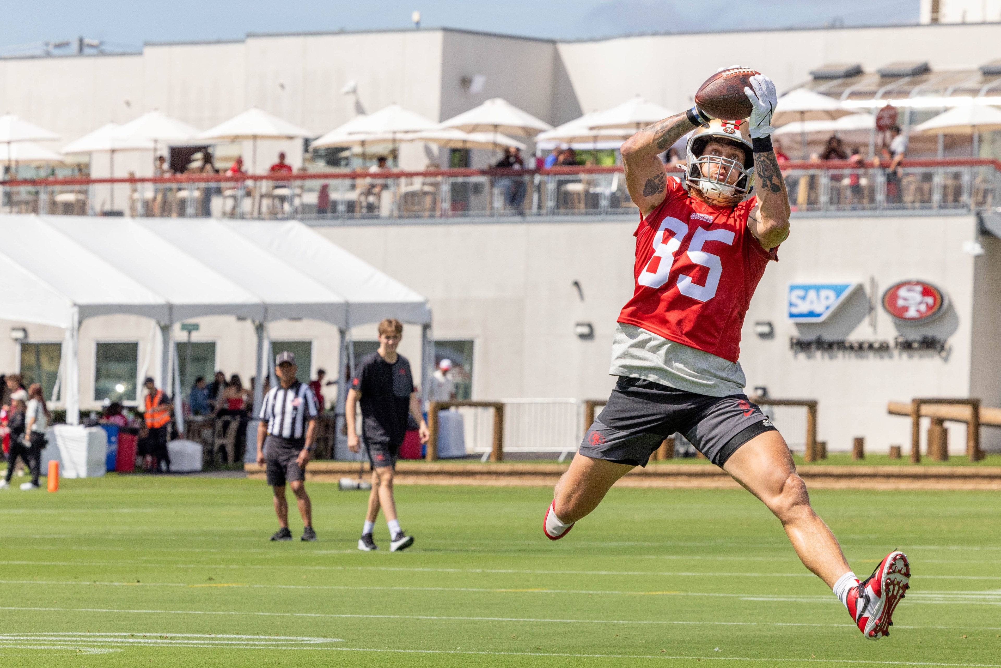 A football player in a red jersey, number 85, leaps to catch a ball during practice on a sunny day, with spectators and tents in the background.