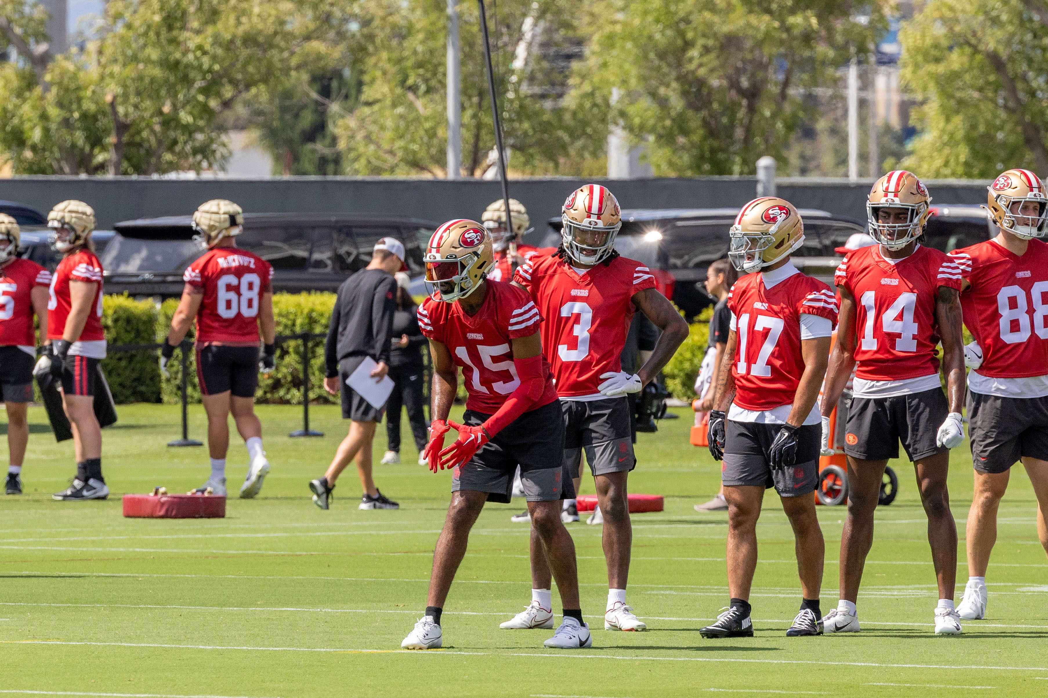 Several football players in red jerseys and helmets stand on a grassy field during practice. They're arranged in a line, with trees and equipment in the background.