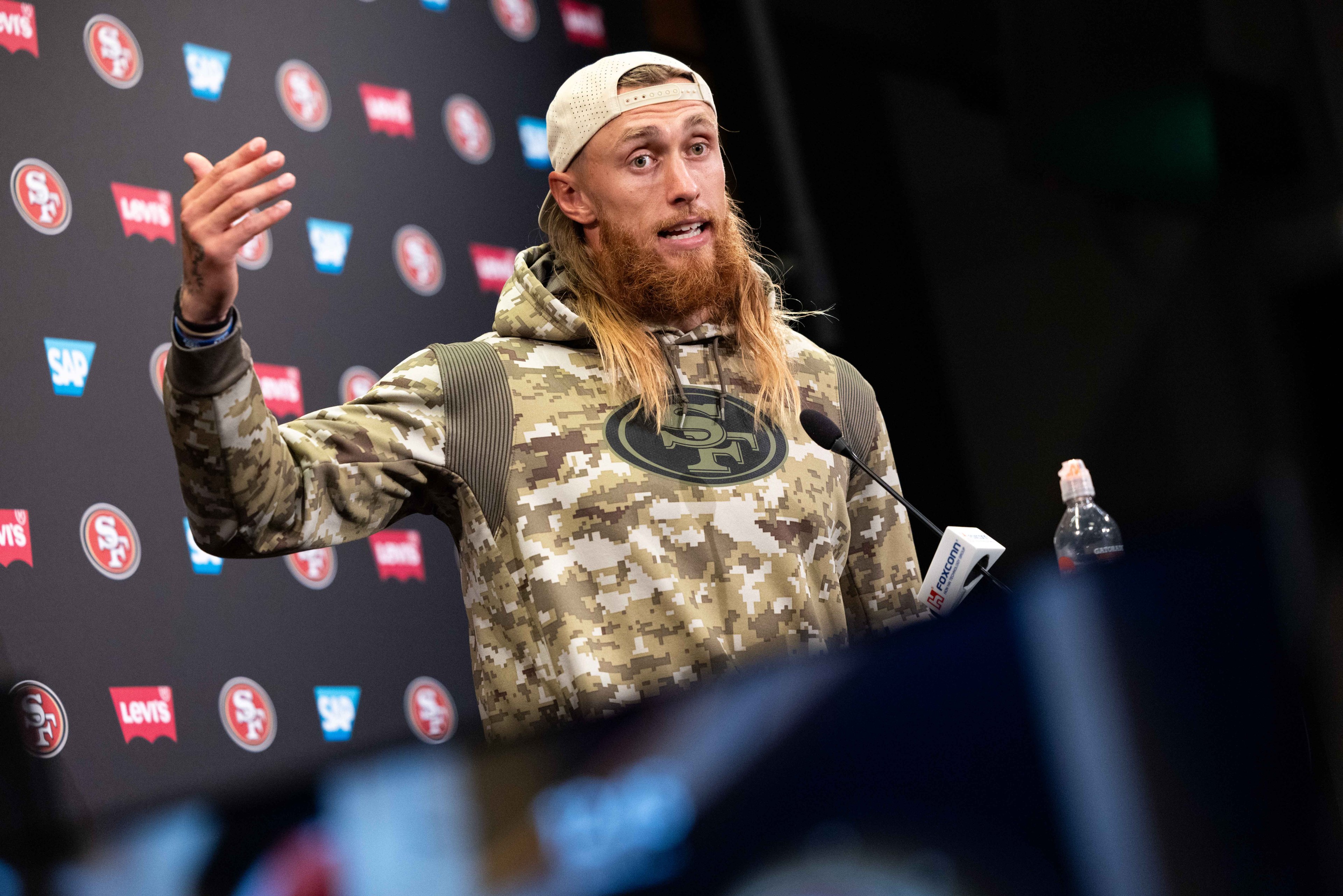 A person in a camouflage hoodie and backward cap is speaking at a podium with microphones. The backdrop features logos of the San Francisco 49ers and sponsors.