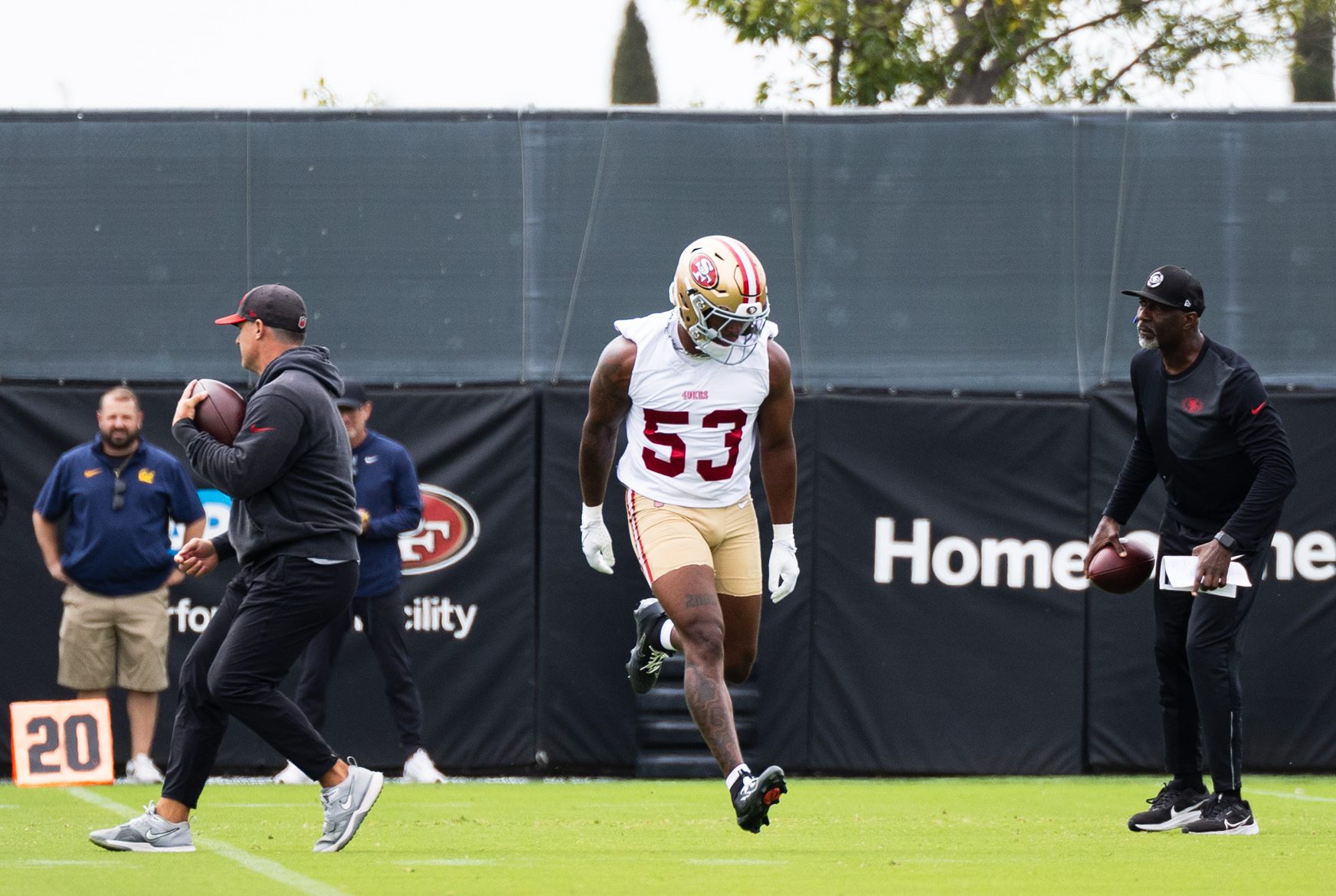 A football practice session shows a player in a 49ers jersey and two coaches. The player is mid-stride, wearing a helmet, while one coach holds a football.