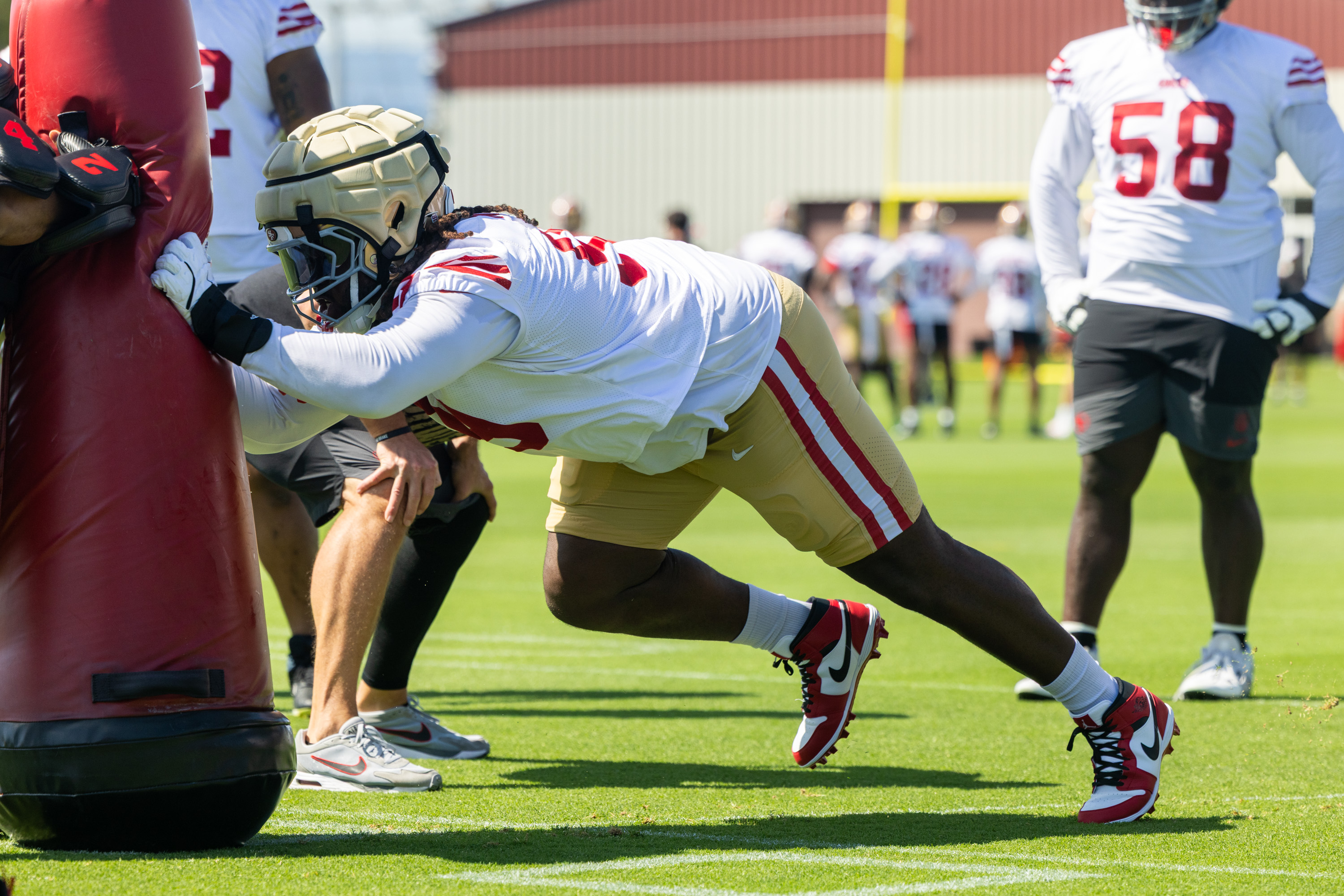 A football player in athletic gear pushes against a padded dummy on a grass field, while another player observes in the background.