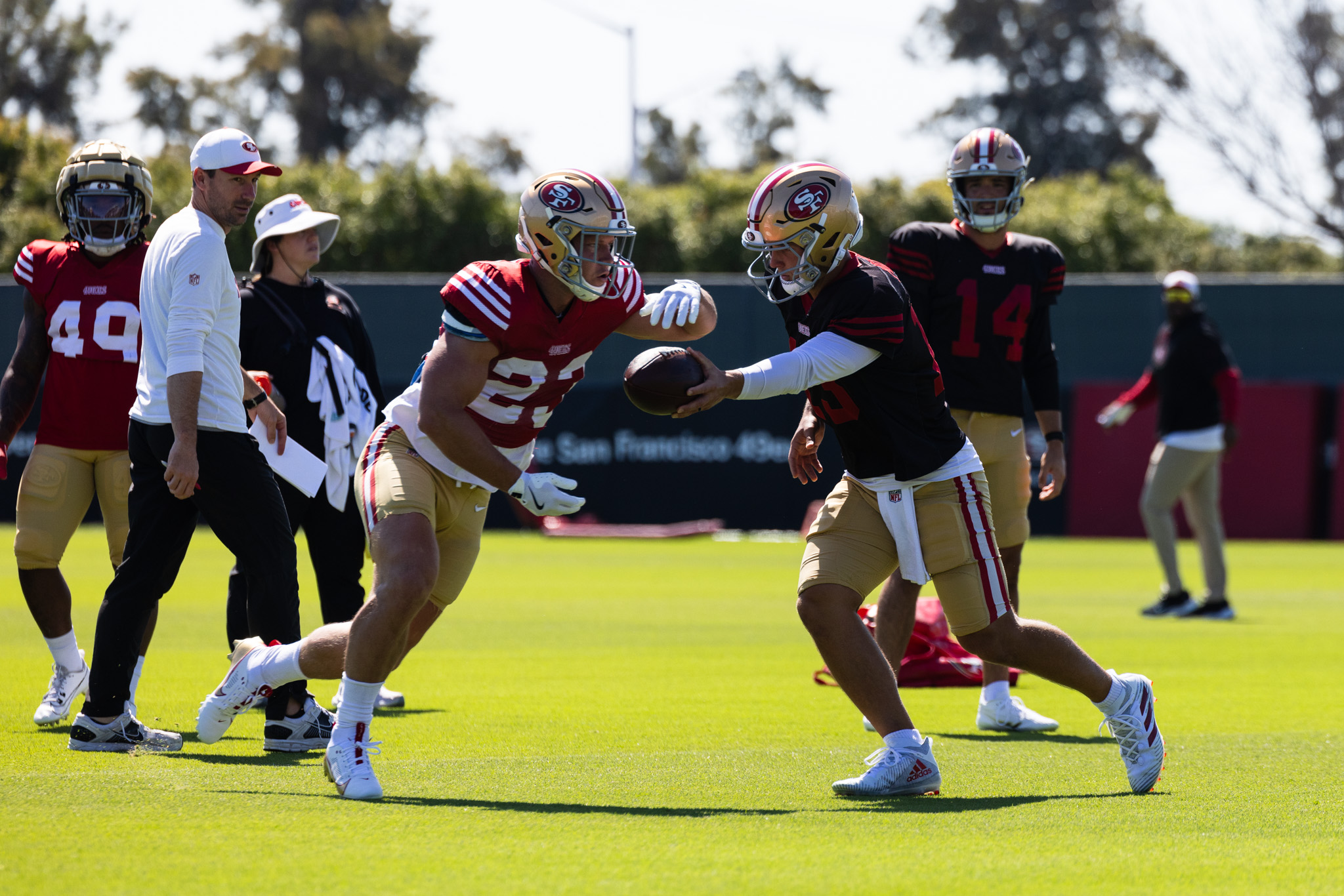 Two football players in red and gold uniforms are practicing a handoff on a grassy field. Coaches and other players are visible in the background.