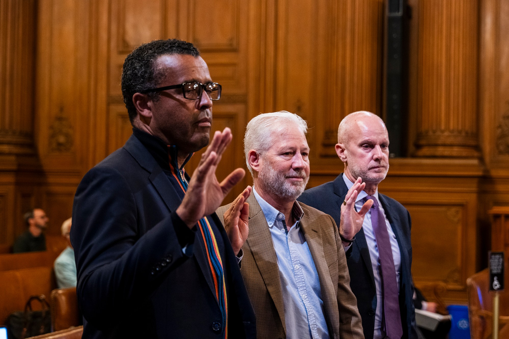 Three men are standing with raised hands in a wood-paneled room, possibly taking an oath. They are wearing formal attire and appear serious.