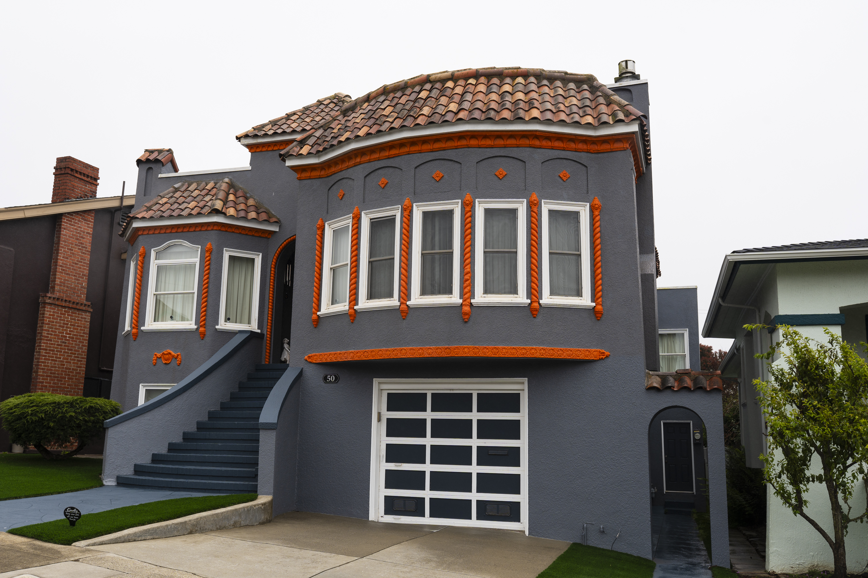 The image shows a gray house with a tile roof and vibrant orange trim. It has arched and rectangular windows, a curved staircase, and a garage.