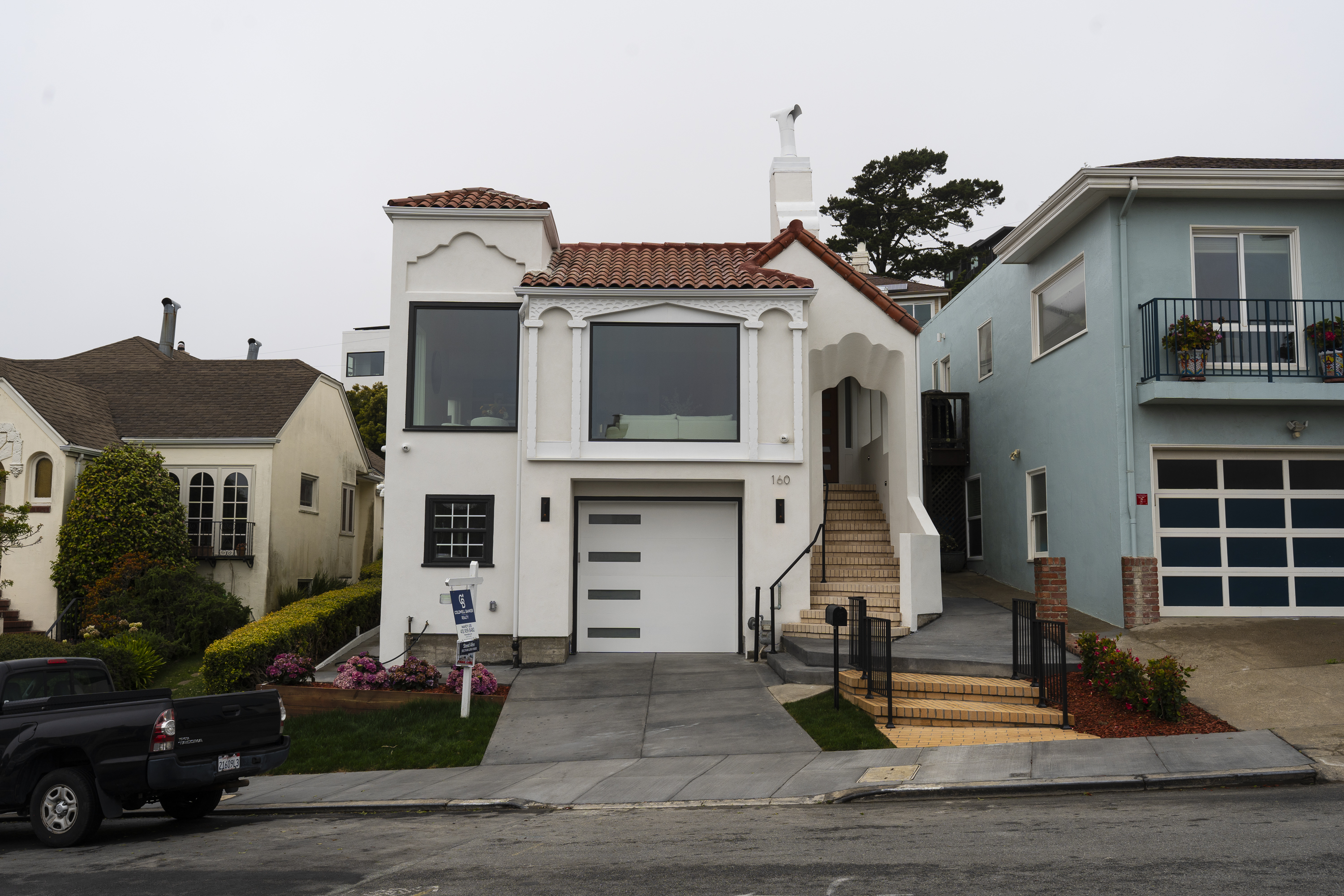 A two-story white house with red roof tiles, a large garage, and arched entryway sits between two homes. A black truck is parked in front.