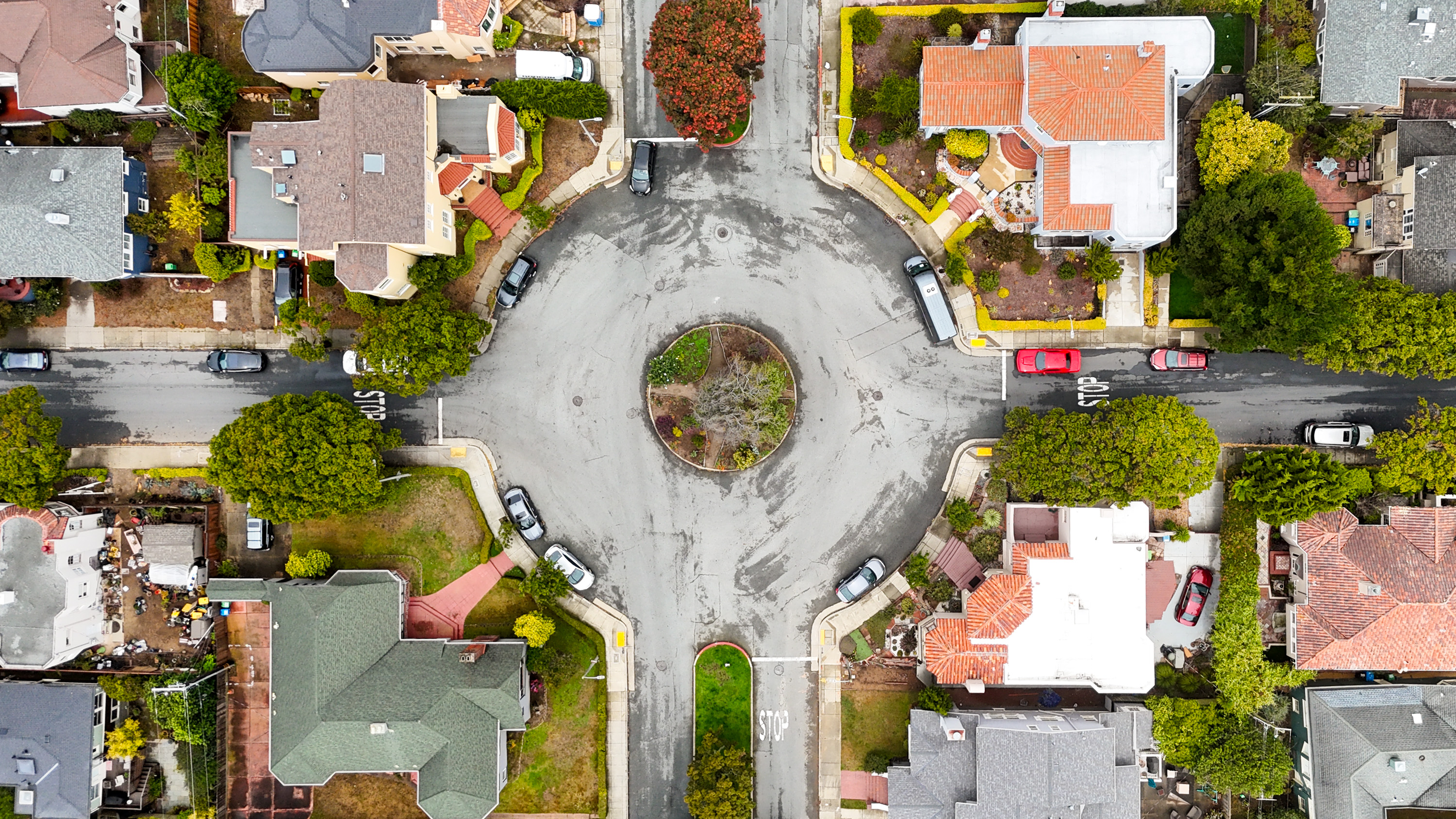 The image shows a roundabout in a suburban neighborhood, surrounded by houses with varied roof designs. Cars are parked along the streets, and greenery is abundant.