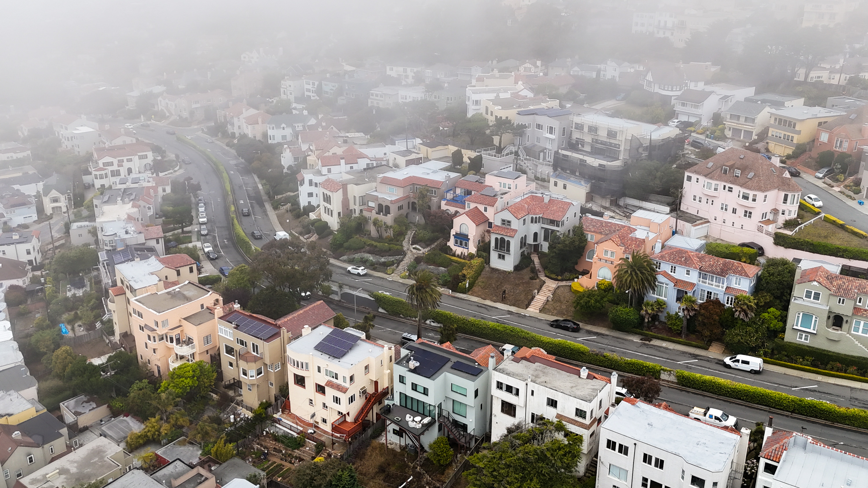 The image shows a foggy aerial view of a hilly residential area with trees. There are winding streets lined with colorful, multi-story houses and greenery.