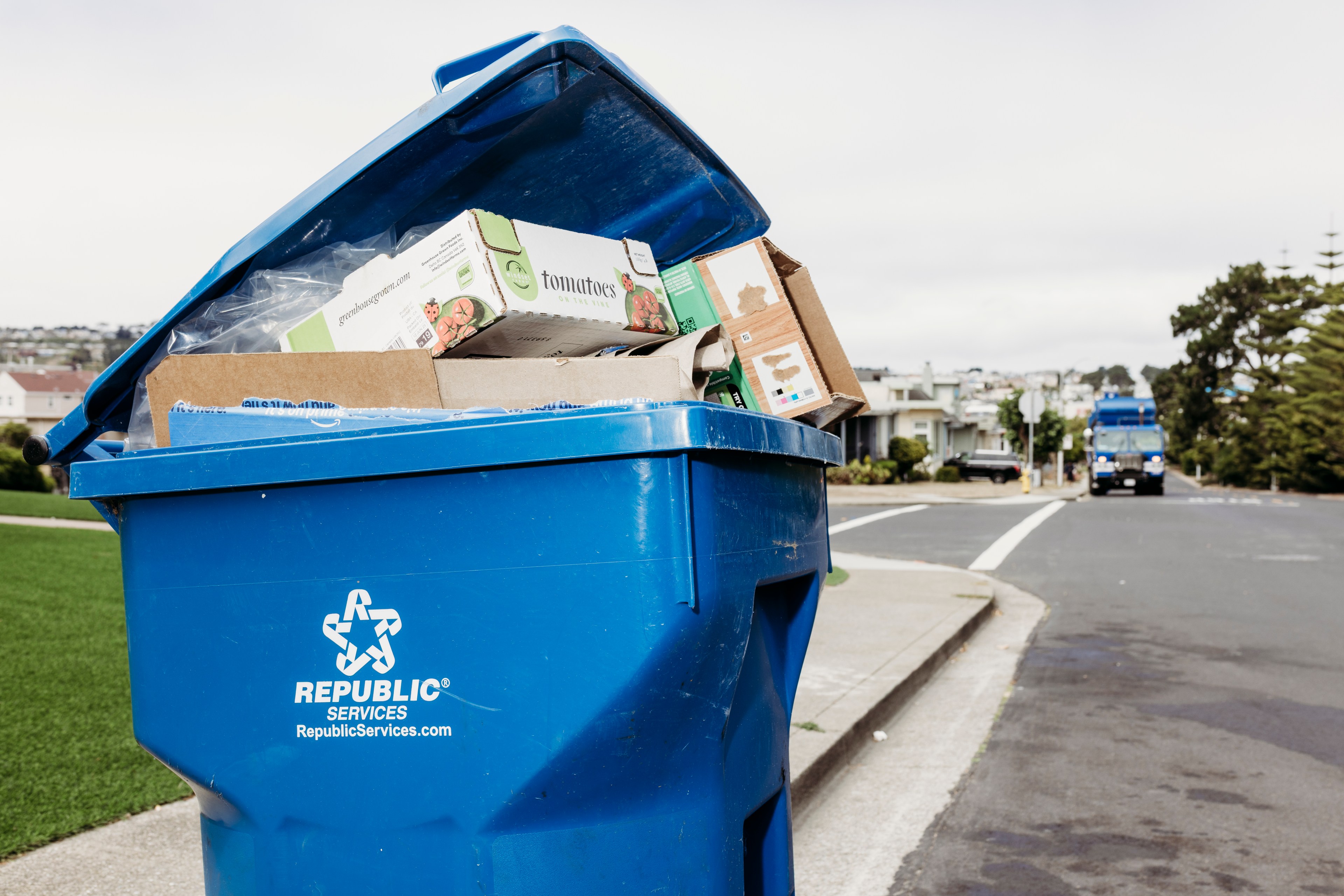 A blue recycling bin is overflowing with cardboard boxes and sits by the roadside. A blue garbage truck approaches in the background on a suburban street.