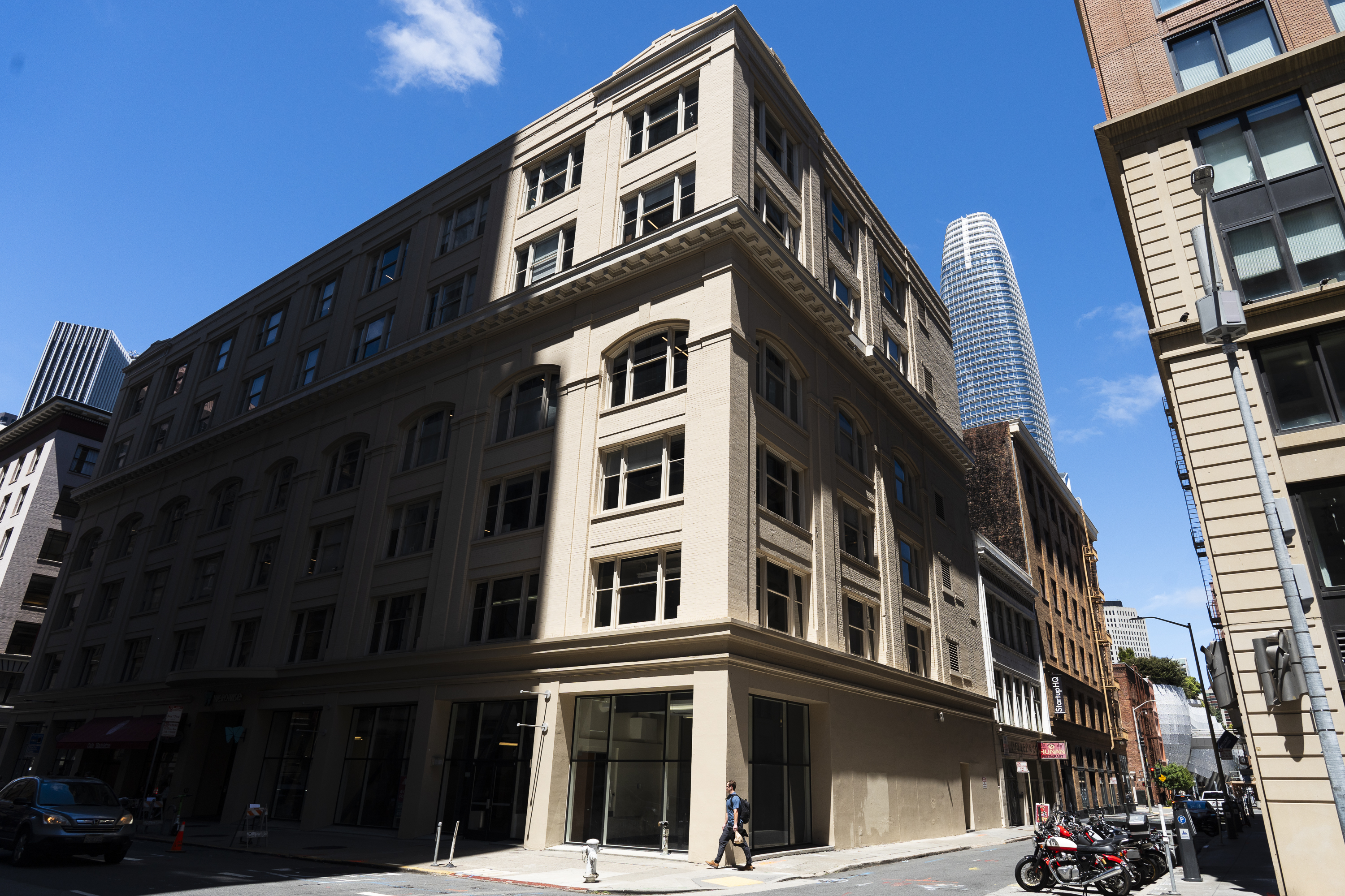A beige, multi-story building stands on a sunny street corner with a blue skyscraper visible in the background. A person walks by, and motorcycles are parked nearby.