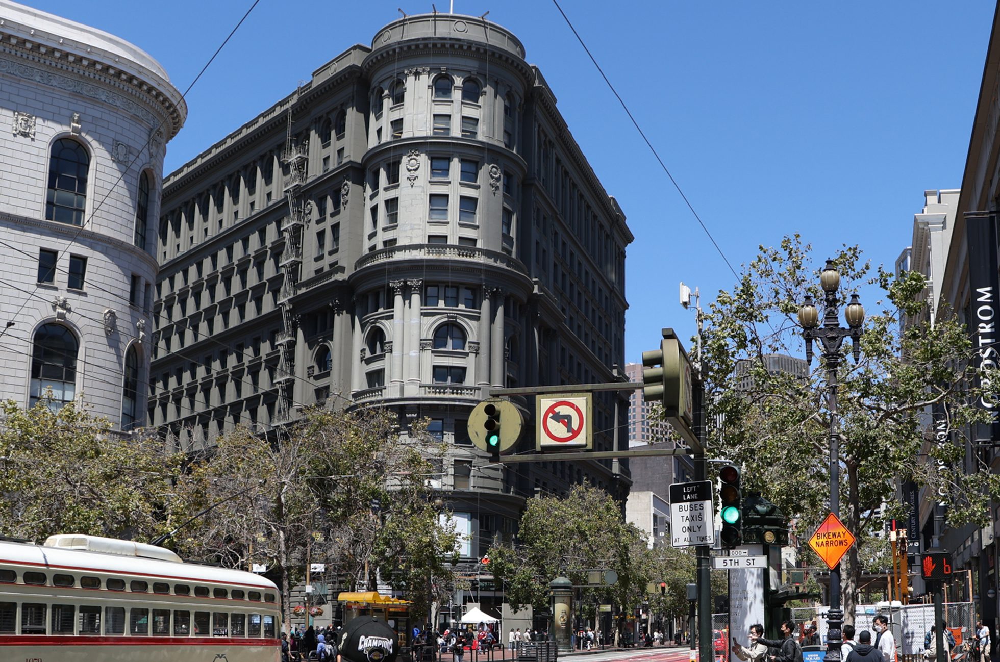A busy urban scene shows historic, ornate buildings and a streetcar, with lush trees lining the streets. A street sign shows no left turns.