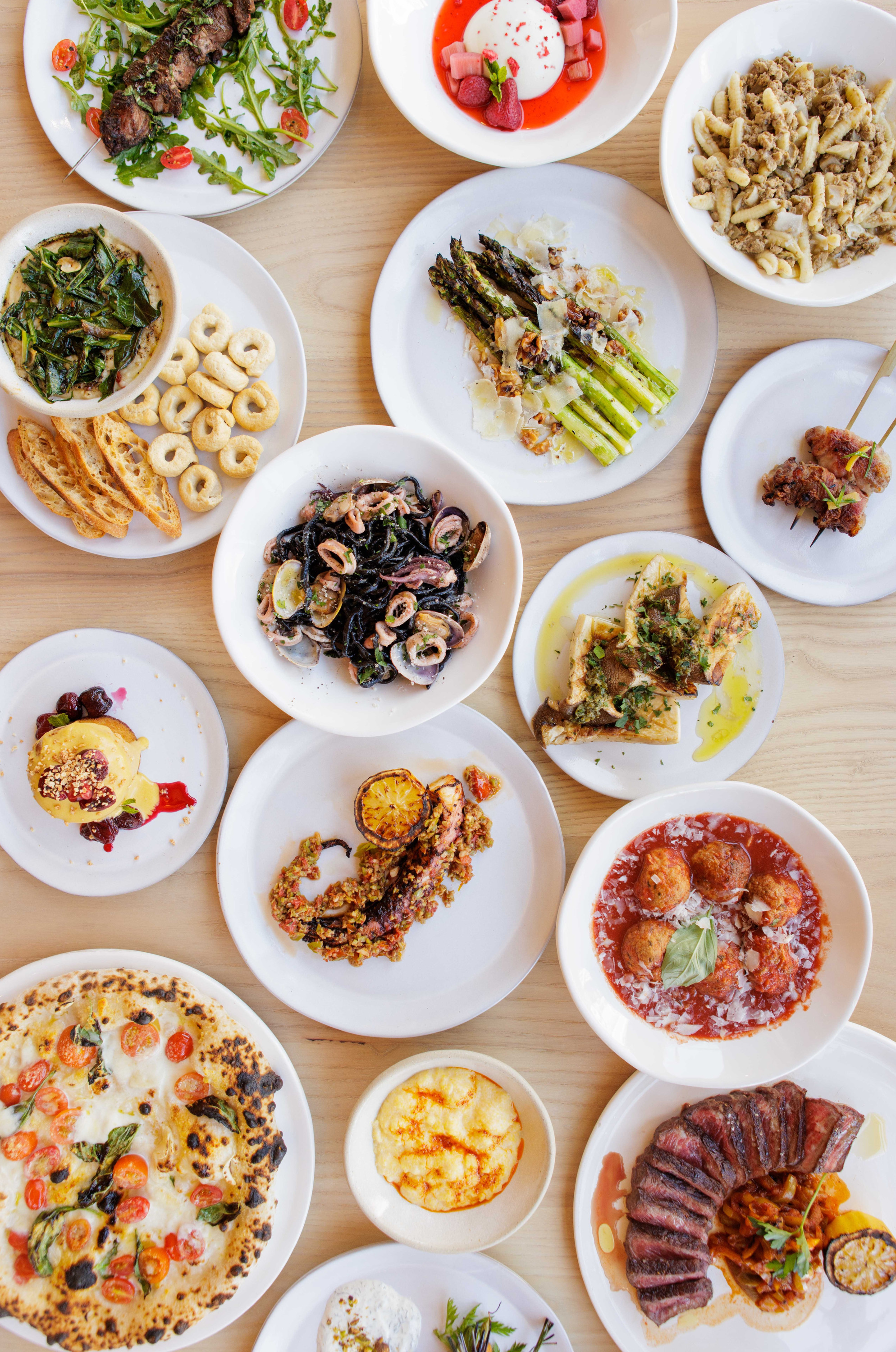 A table is filled with various Italian dishes, including pasta, pizza, seafood, skewers, and desserts, all artfully plated on white dishes.