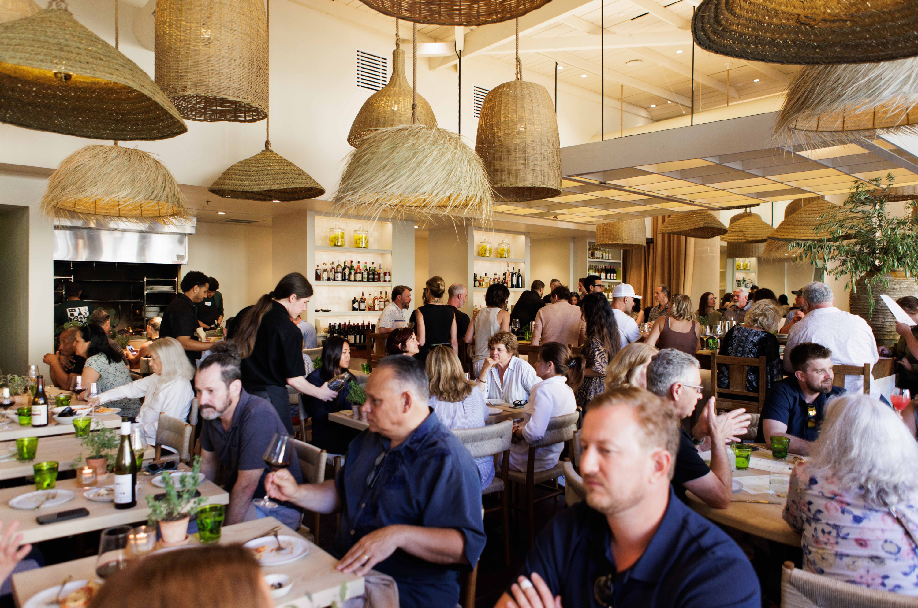 The image shows a bustling restaurant with people dining and chatting at tables. Woven basket-like lights hang from the ceiling, and a bar is visible in the background.
