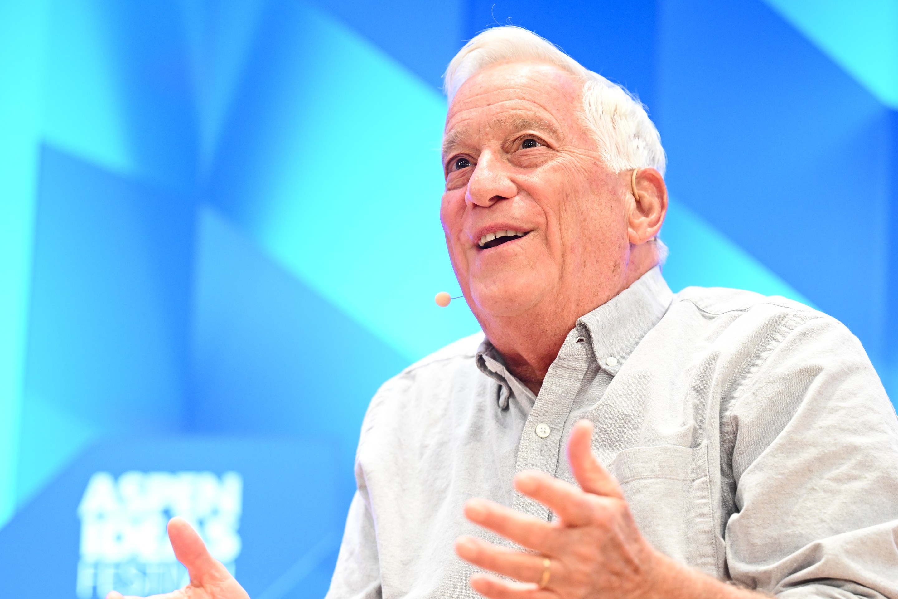 An older man with white hair is speaking animatedly on stage, wearing a light gray shirt. He's gesturing with his hands; a microphone is visible on his cheek.