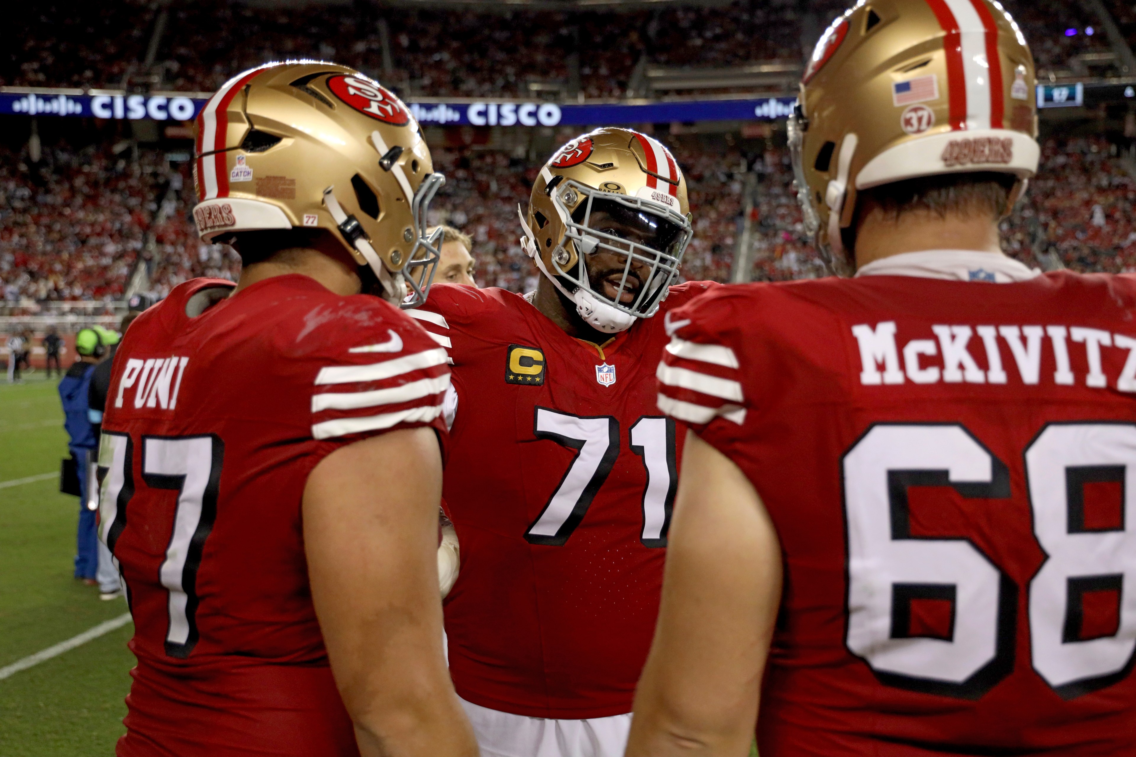 Three football players wearing red 49ers uniforms and helmets stand in a huddle on the field, with a crowded stadium in the background.