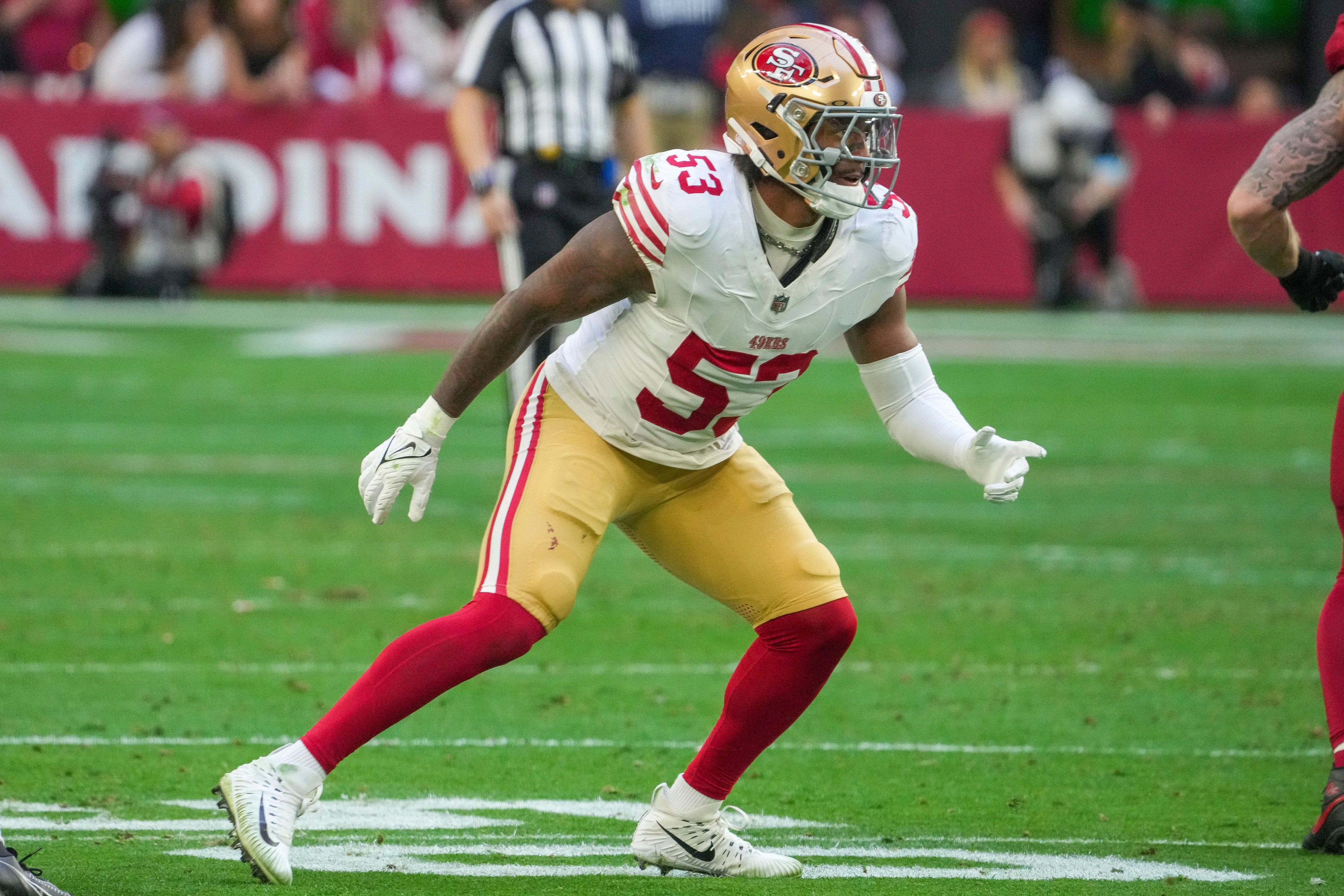 A football player wearing a white 49ers jersey and gold helmet is in an action stance on a grass field. He's wearing red leggings and white cleats.