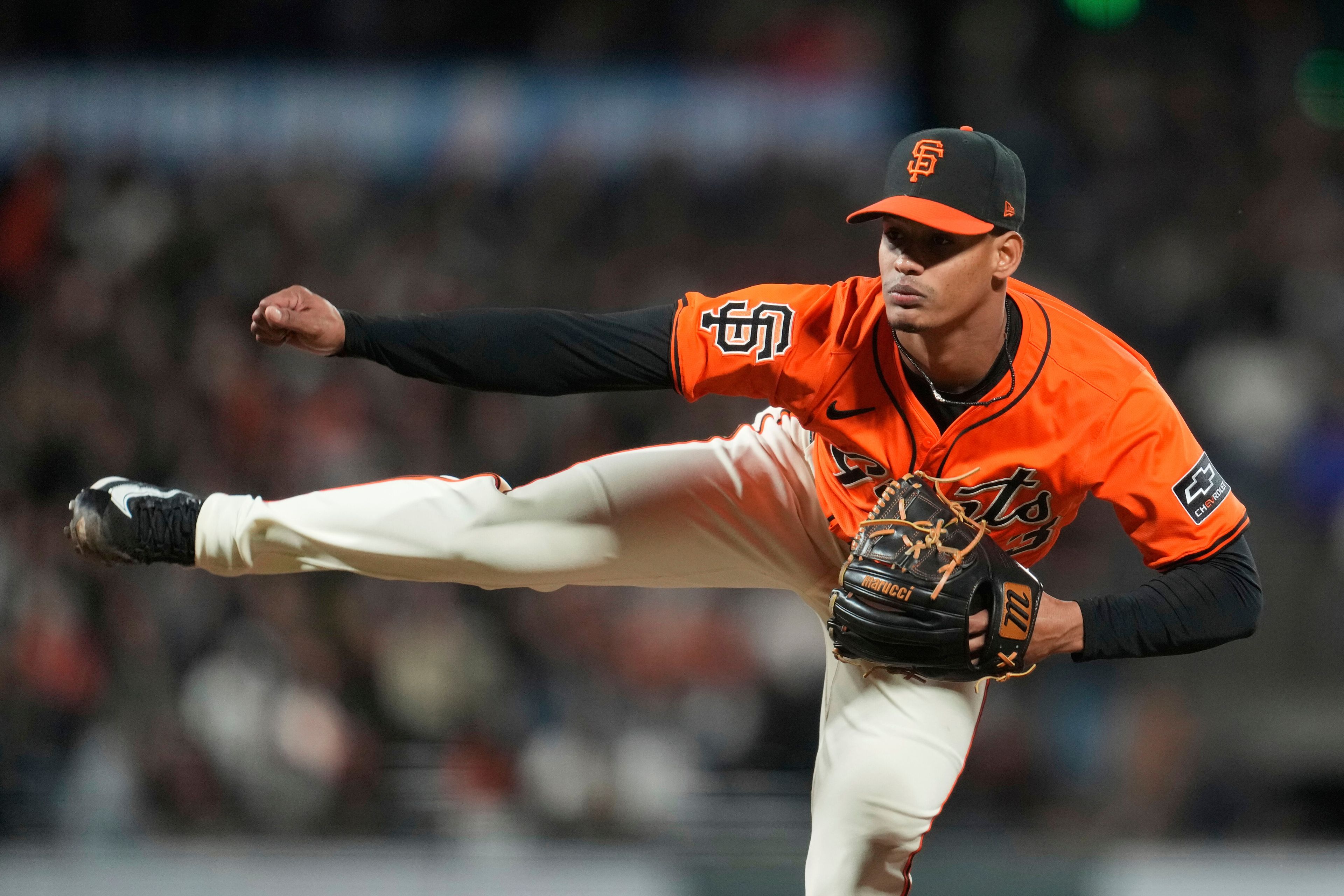 A baseball pitcher in an orange jersey with "SF" on the cap throws a pitch, with one leg high in the air. His glove is positioned near his chest.