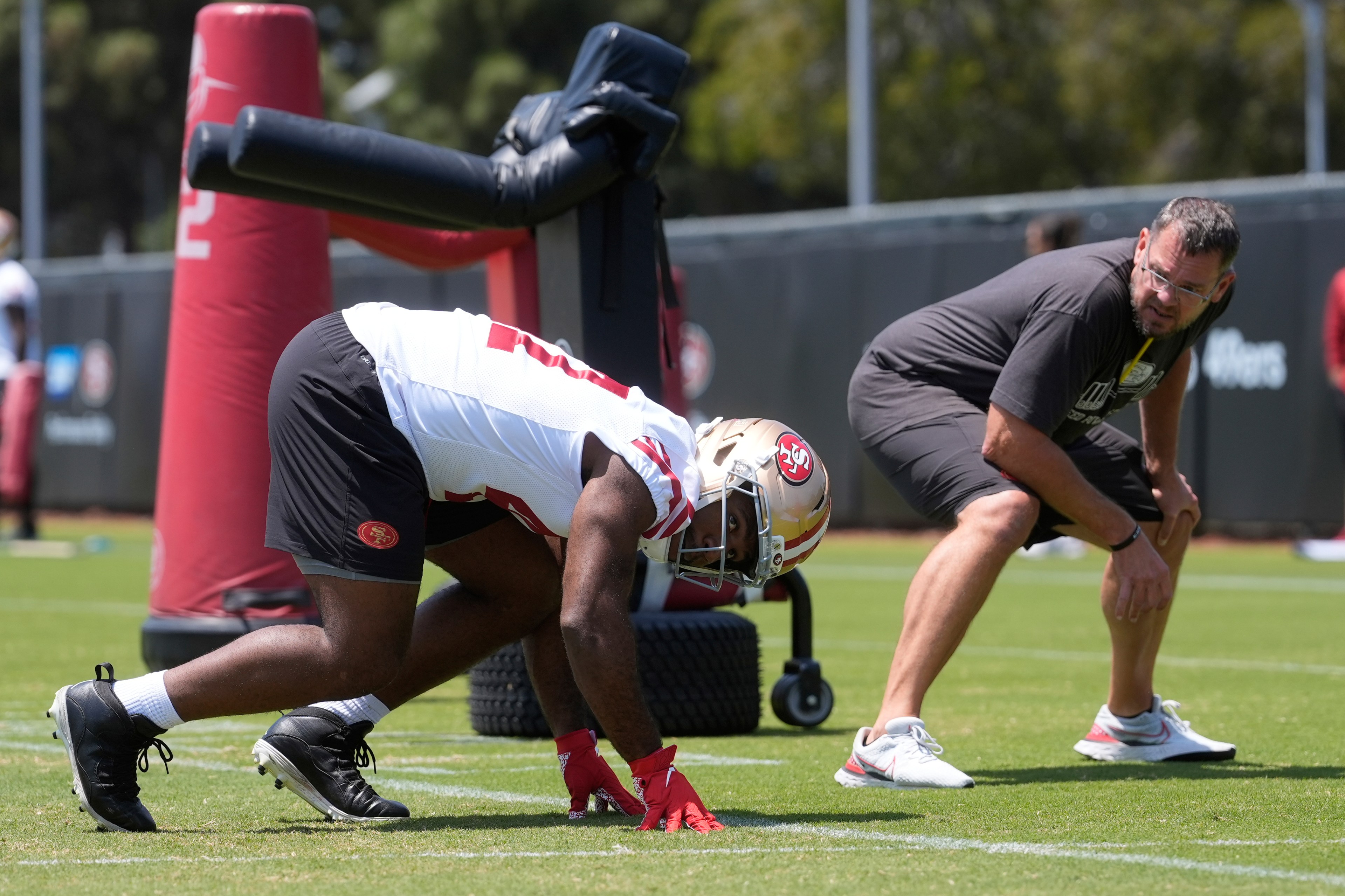 A football player in full gear, wearing a helmet, crouches in a ready stance on a grassy field. A coach observes nearby, both focused on practice.