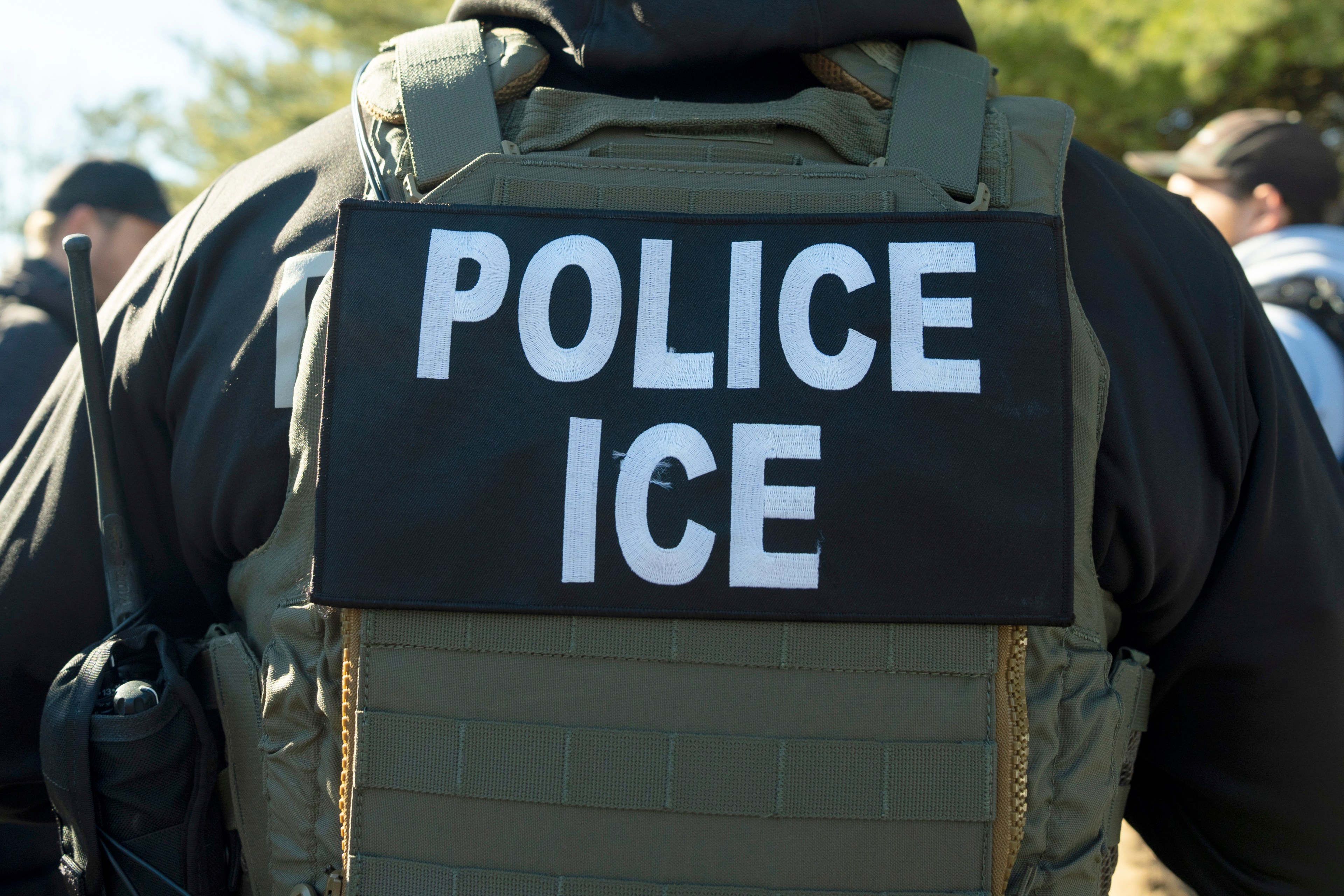 A person is wearing a tactical vest with a patch labeled "POLICE ICE." The vest is dark, and there are others in the background, possibly outdoors.