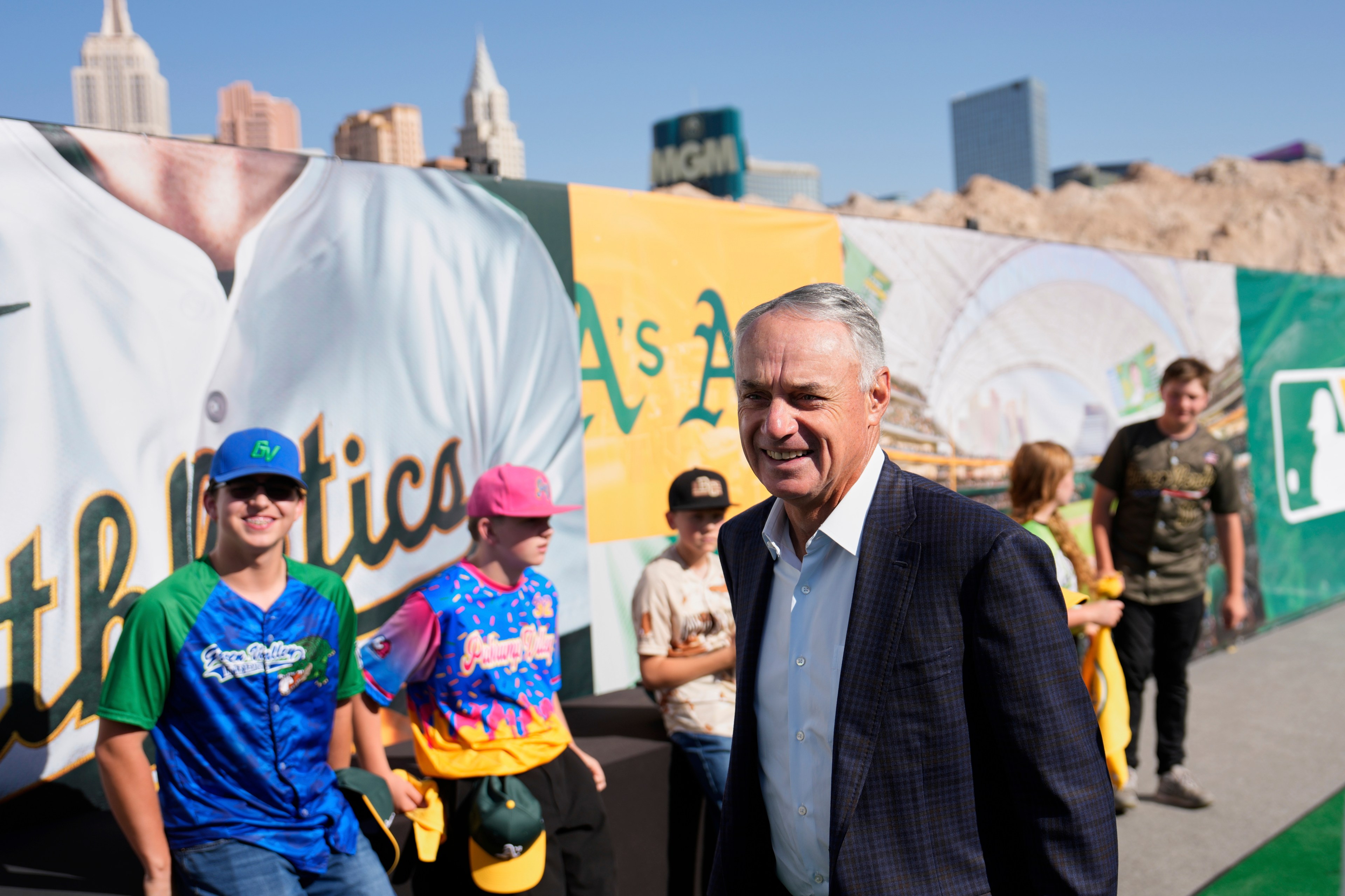 A man in a suit smiles while standing near a colorful baseball-themed backdrop. Behind him, a group of people in baseball jerseys and caps are gathered.