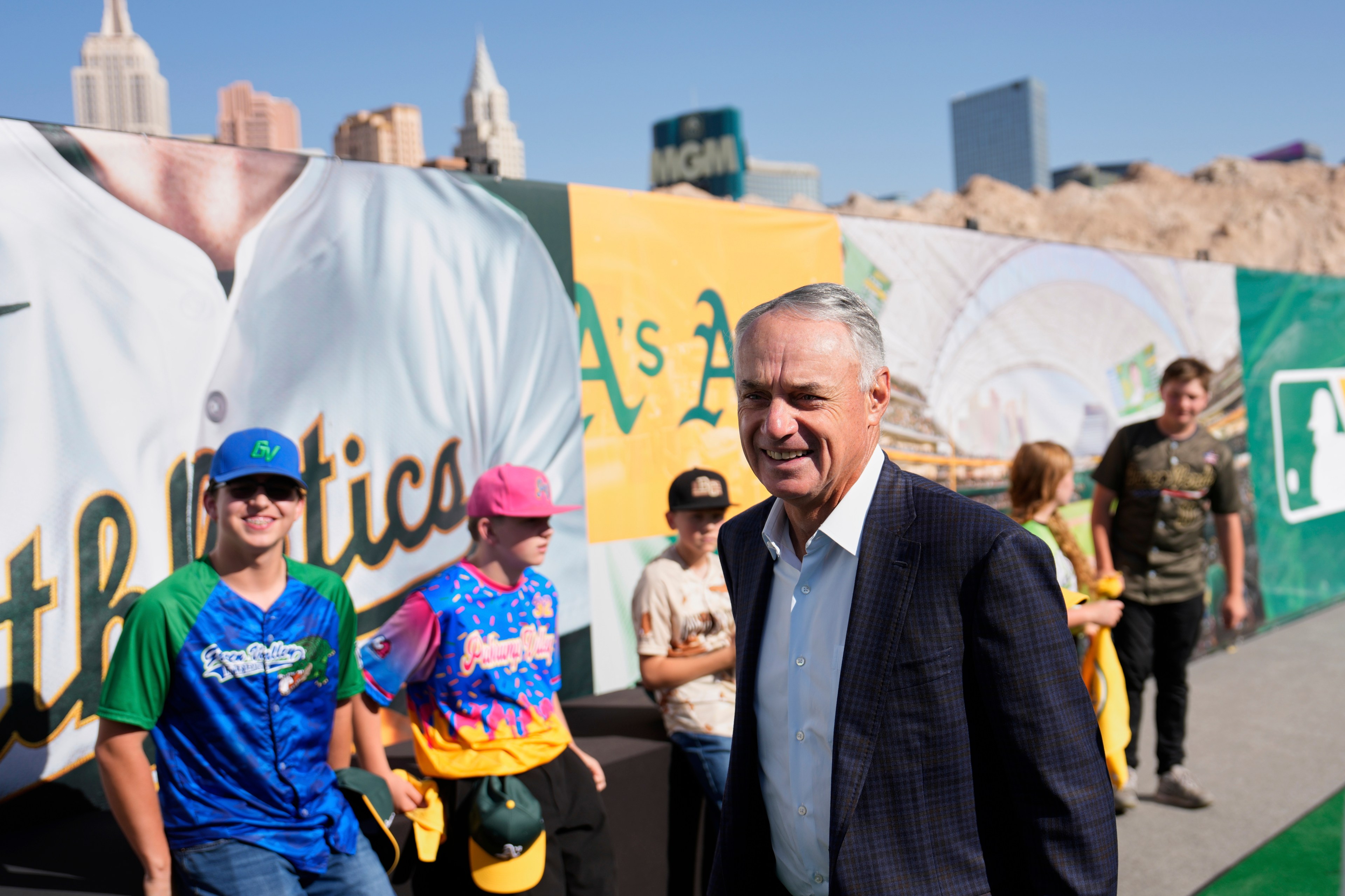 A man in a suit smiles while standing near a colorful baseball-themed backdrop. Behind him, a group of people in baseball jerseys and caps are gathered.