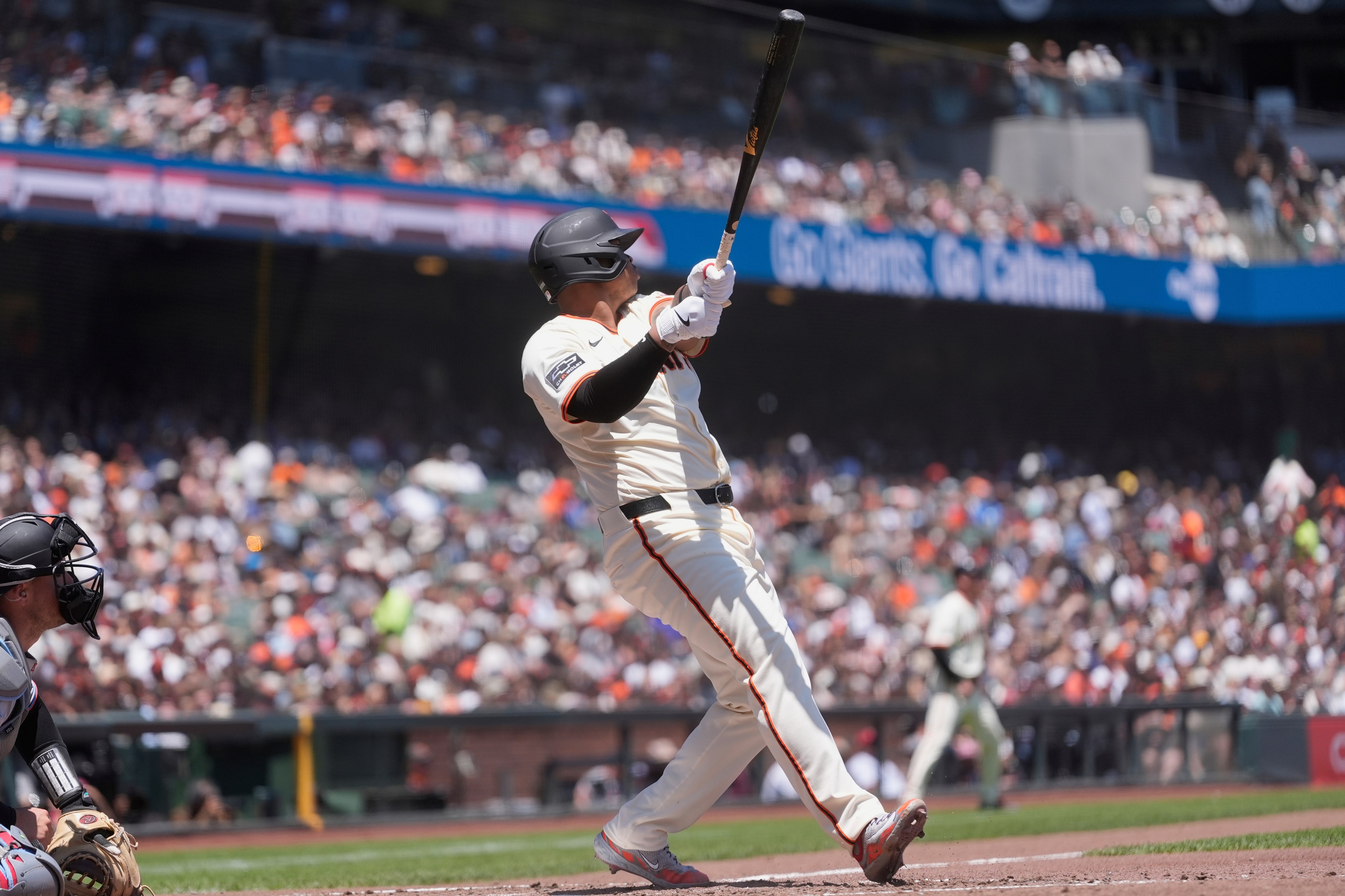 A baseball player, wearing a white uniform and black helmet, swings a bat at a stadium. A catcher crouches nearby and a large crowd fills the stands.