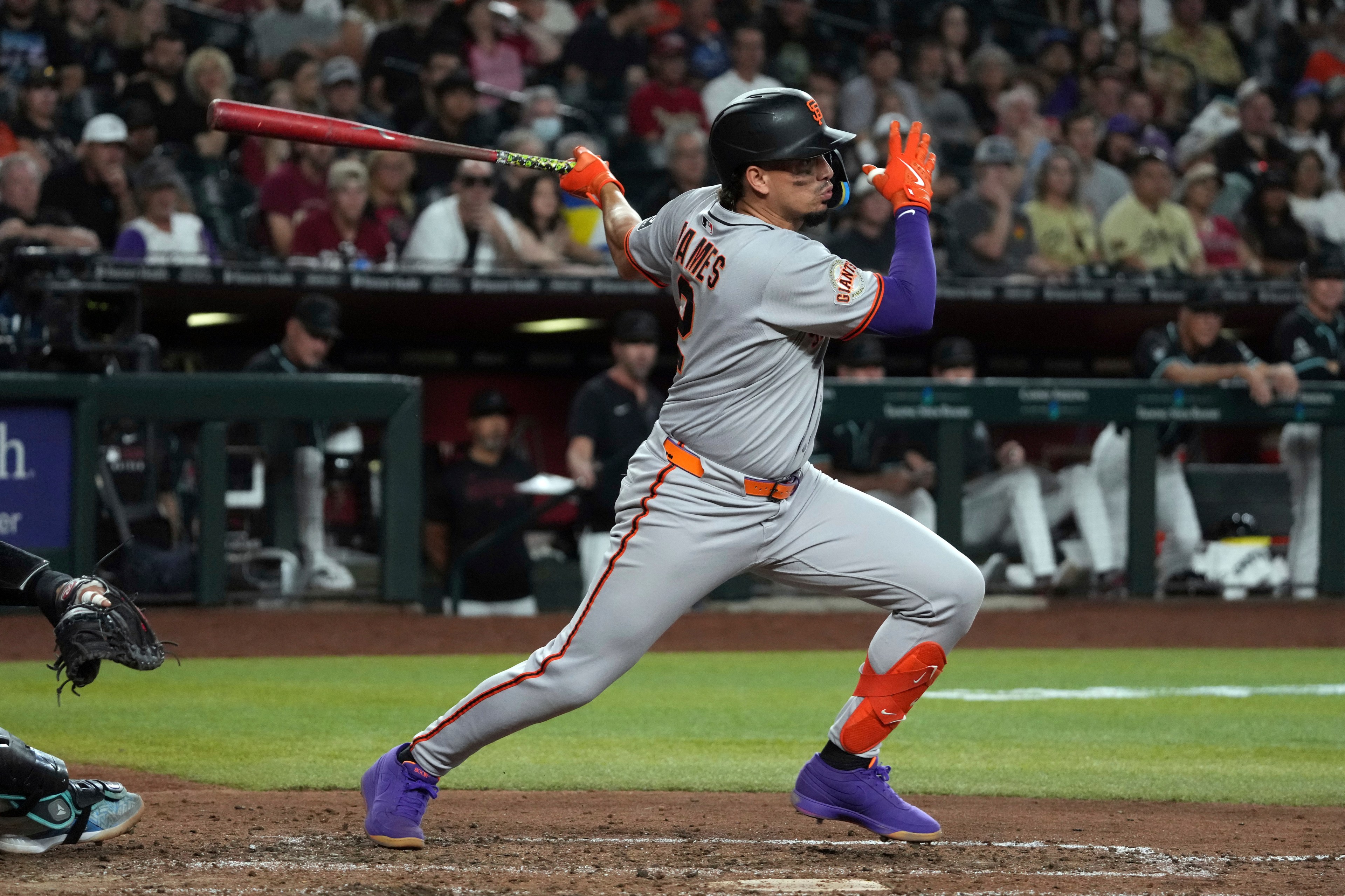 A baseball player in a gray uniform swings a bat during a game. He wears bright gloves and shoes, with an audience in the blurred background.