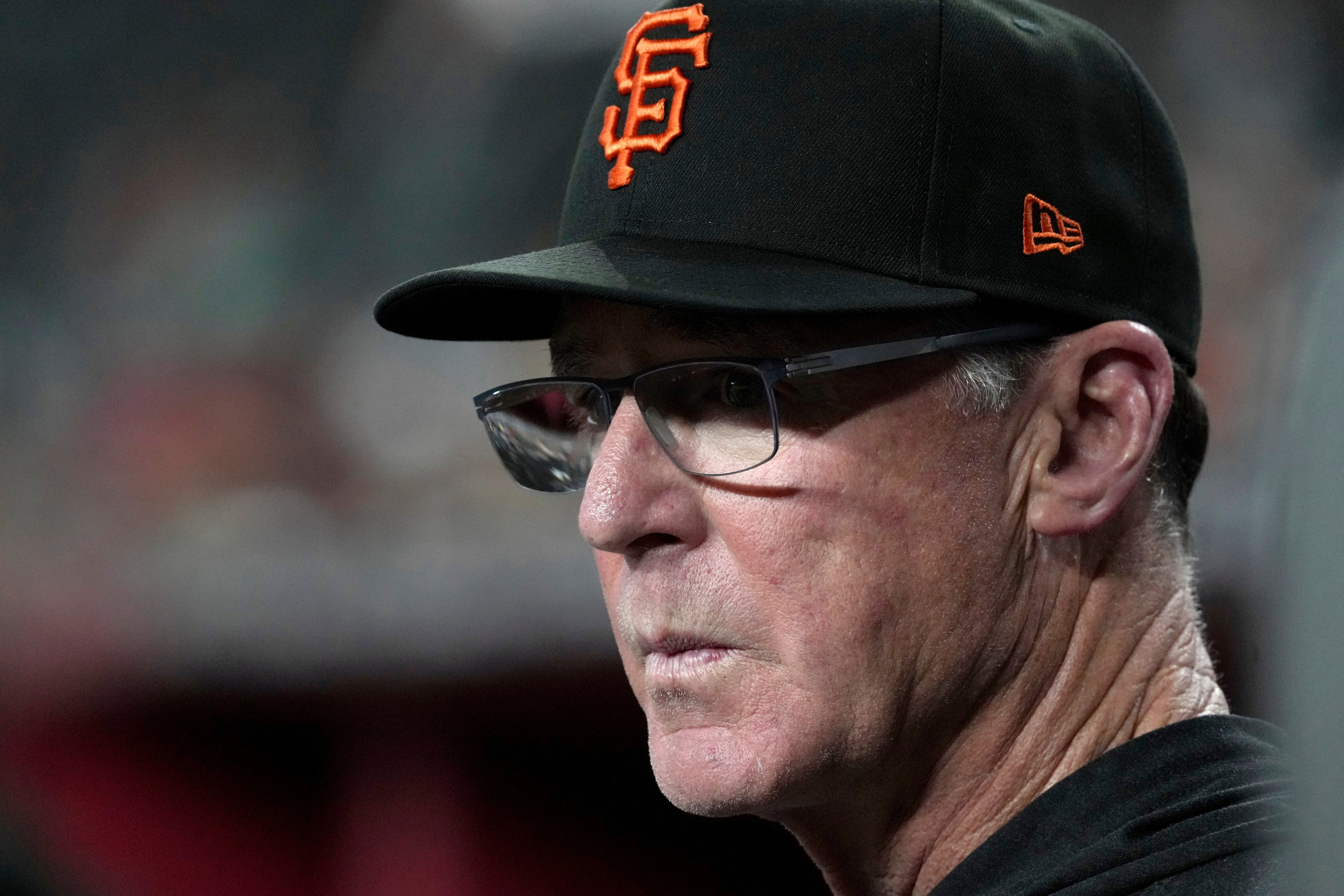 An older man wears glasses and a black baseball cap with an orange &quot;SF&quot; logo. His expression is focused, with a blurred background behind him.