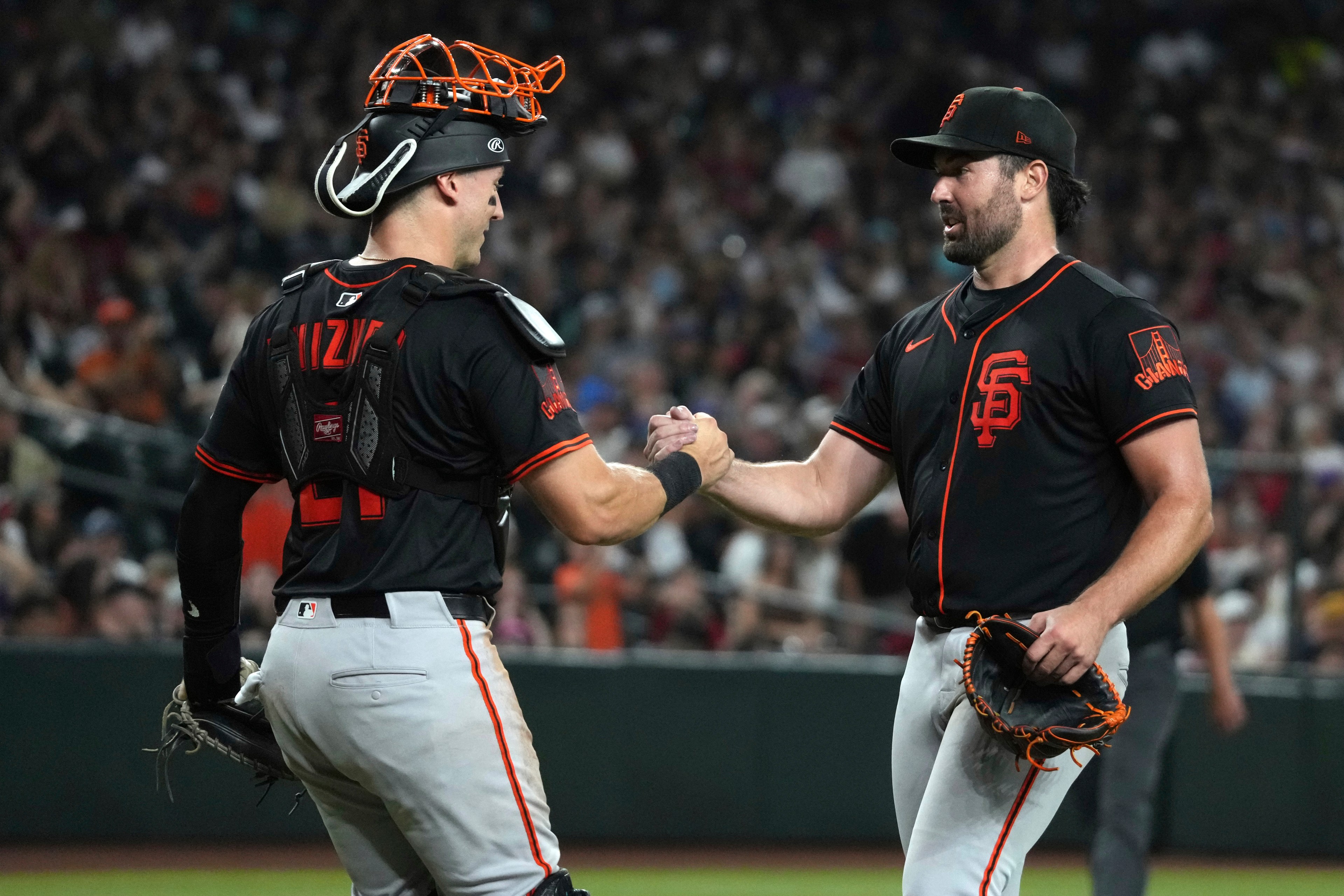 Two baseball players in black uniforms are shaking hands on the field. One wears catcher's gear, the other holds a glove. The background is filled with spectators.
