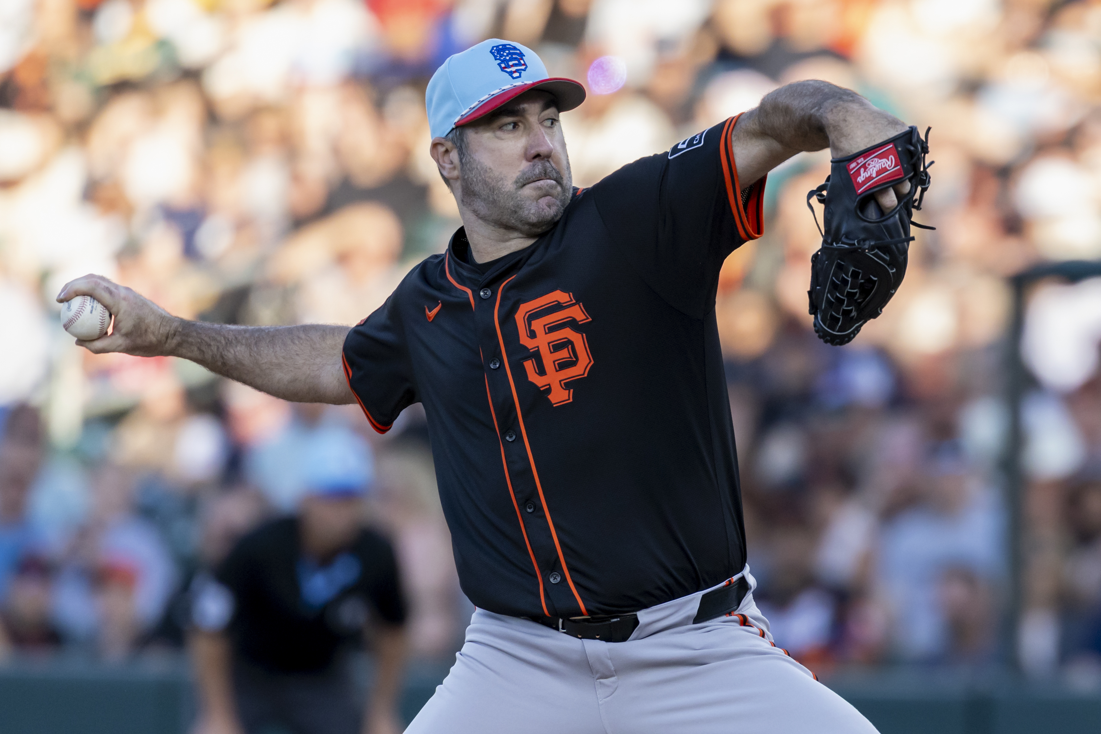 A baseball player in a black jersey with an orange "SF" emblem is mid-pitch on a field. He wears a light blue cap, glove, and holds a baseball. The crowd is blurred behind him.