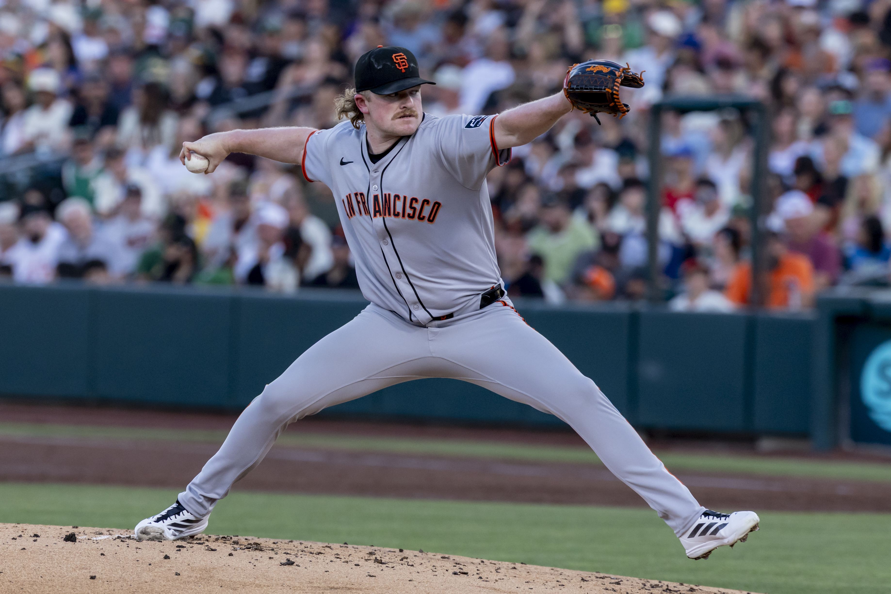 A baseball player in a grey "San Francisco" uniform pitches on a mound with a large crowd in the background. He's mid-throw, with one leg raised.