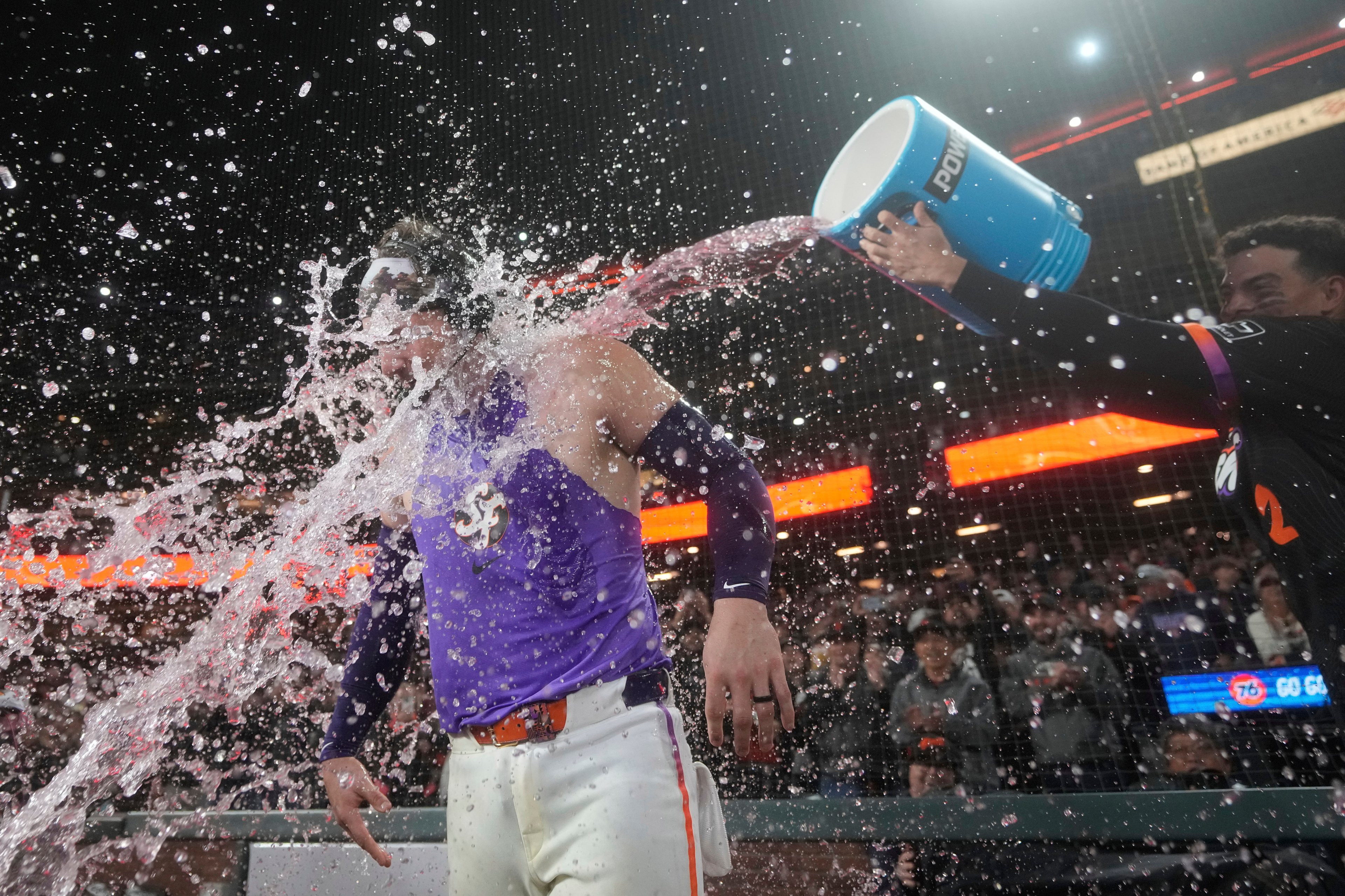 A baseball player in a purple jersey is doused with water from a cooler by a teammate. The water splashes dramatically, and a crowd is visible in the background.