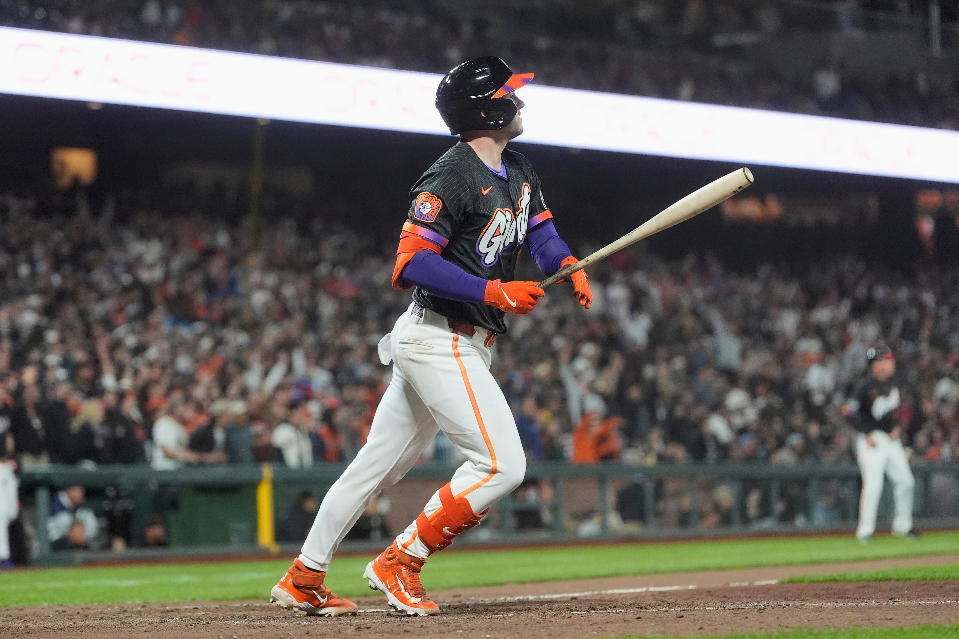 A baseball player in a black and orange uniform watches the ball after hitting it, standing on a field with a cheering crowd in the background.