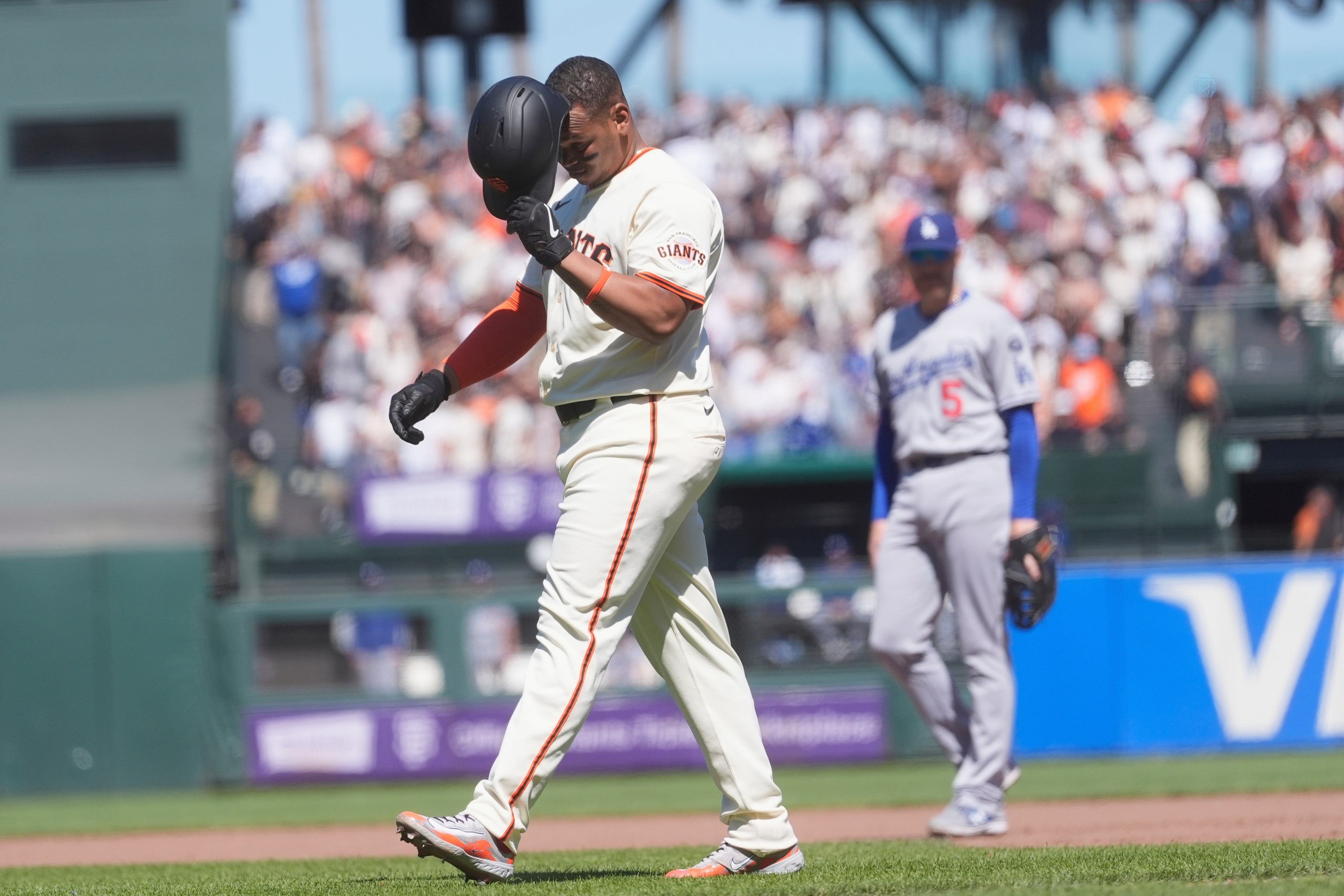 A baseball player in a Giants uniform walks on the field, holding his helmet. A player in a gray Dodgers uniform stands in the background. A crowd is watching.