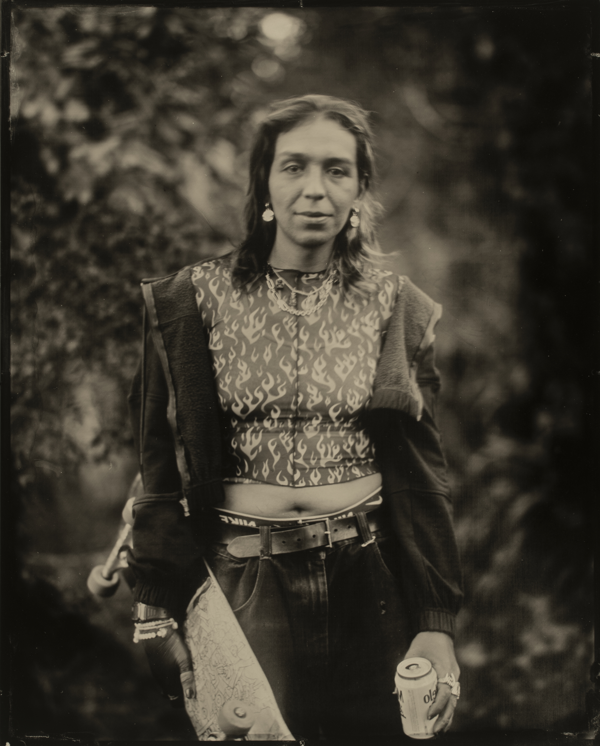 A person with long hair stands outdoors, wearing a flame-patterned shirt, a jacket, and accessories. They hold a drink can and a skateboard adorned with designs.