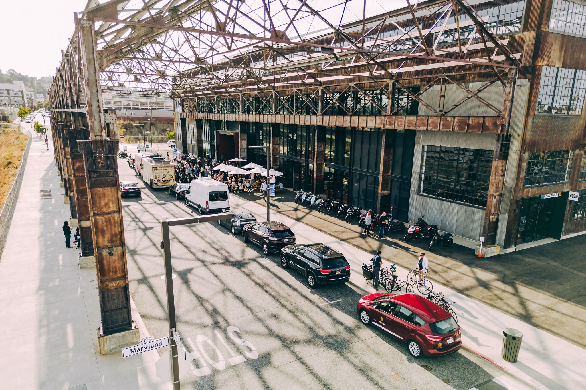 The image shows a busy street next to an industrial-style building with cars, a van, and people walking. Bicycles and motorcycles are lined up outside.