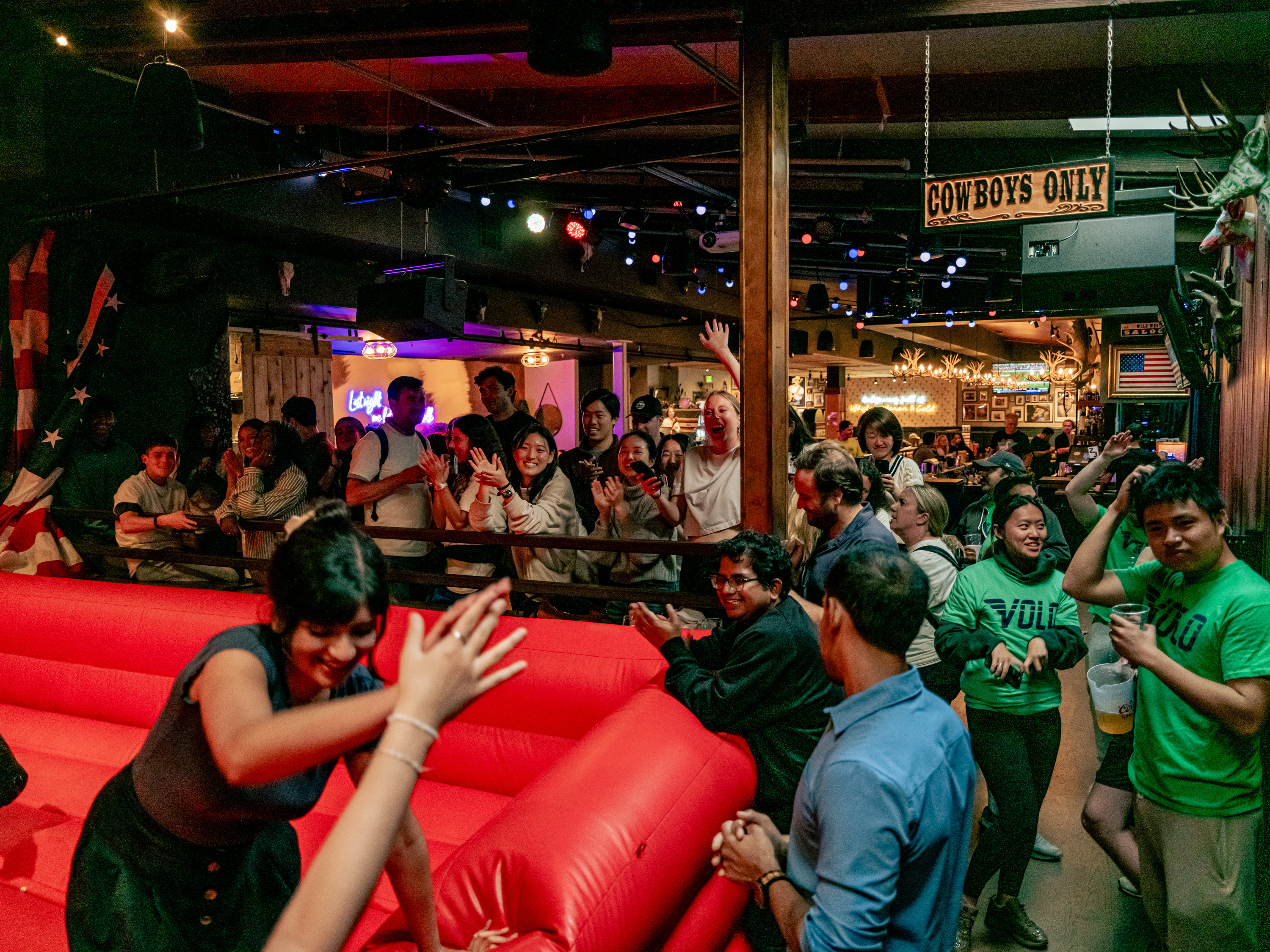 The image shows a lively bar scene with a crowd cheering near a mechanical bull. People are smiling and clapping, while a woman leans on the inflatable.
