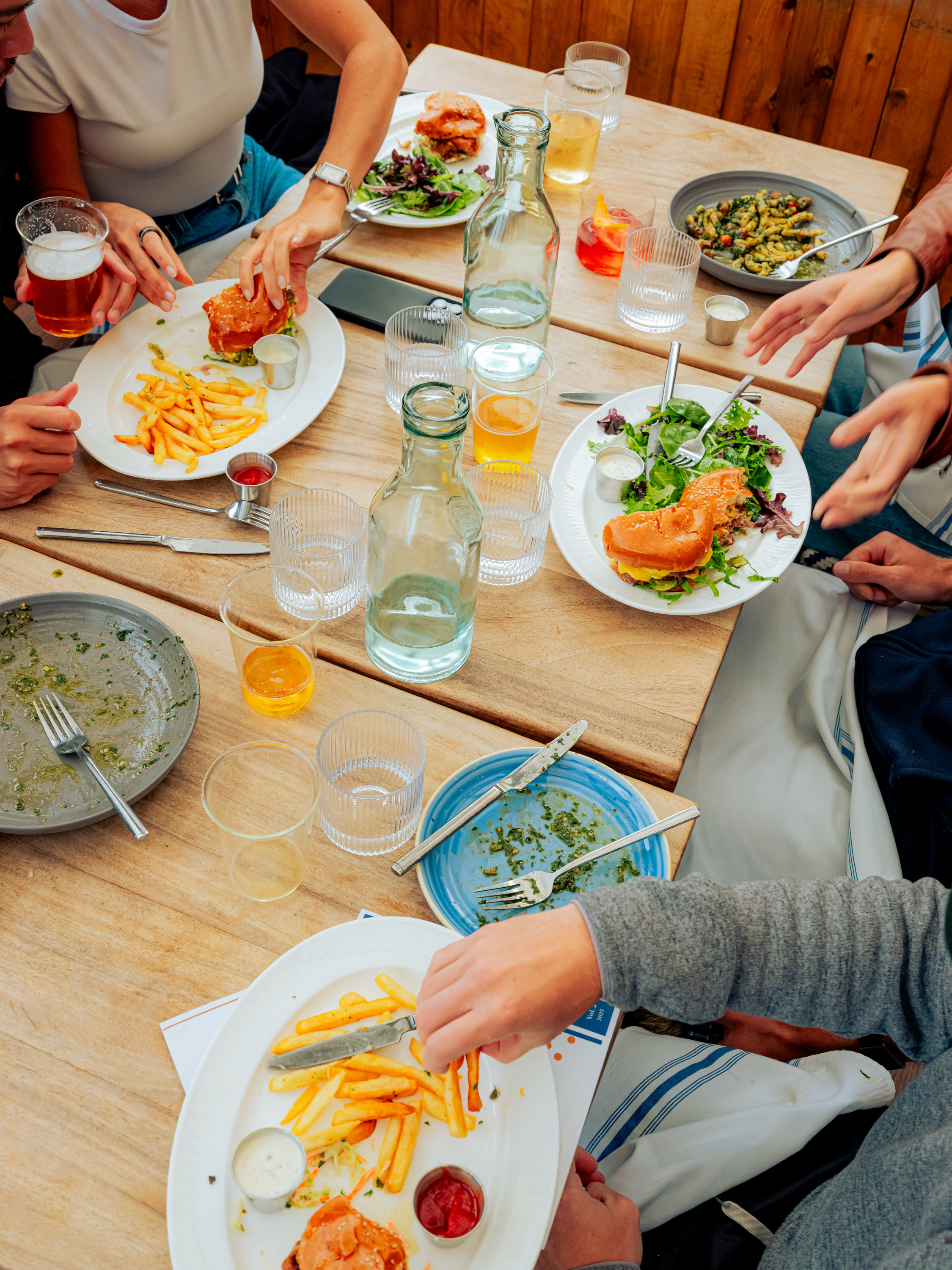 People are enjoying a meal at a wooden table, featuring burgers, fries, salads, drinks, and empty plates. Hands are reaching for food and beverages.