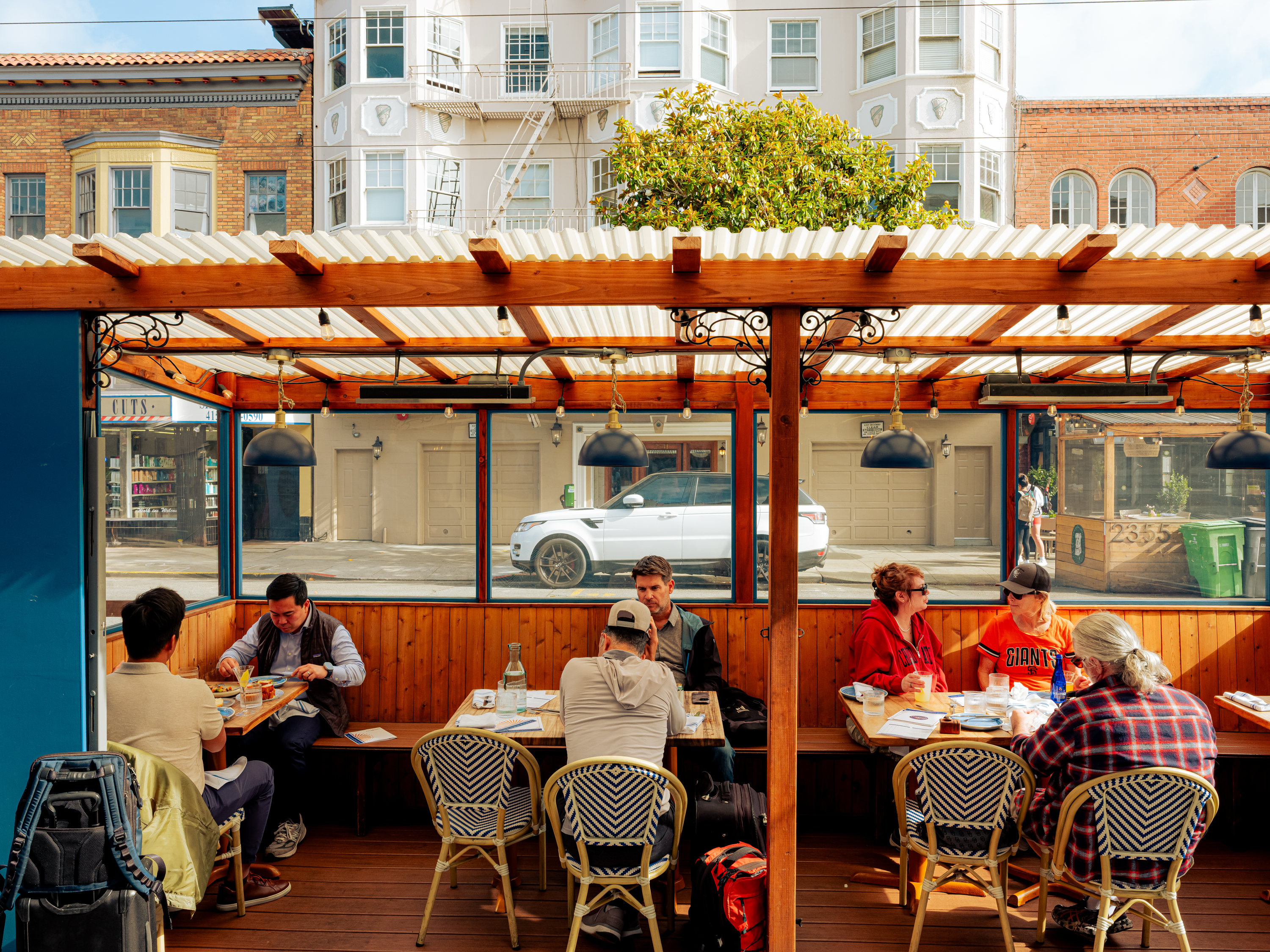 Several people sit at wooden tables under a pergola at an outdoor cafe. The street and buildings are visible in the background through clear panels.