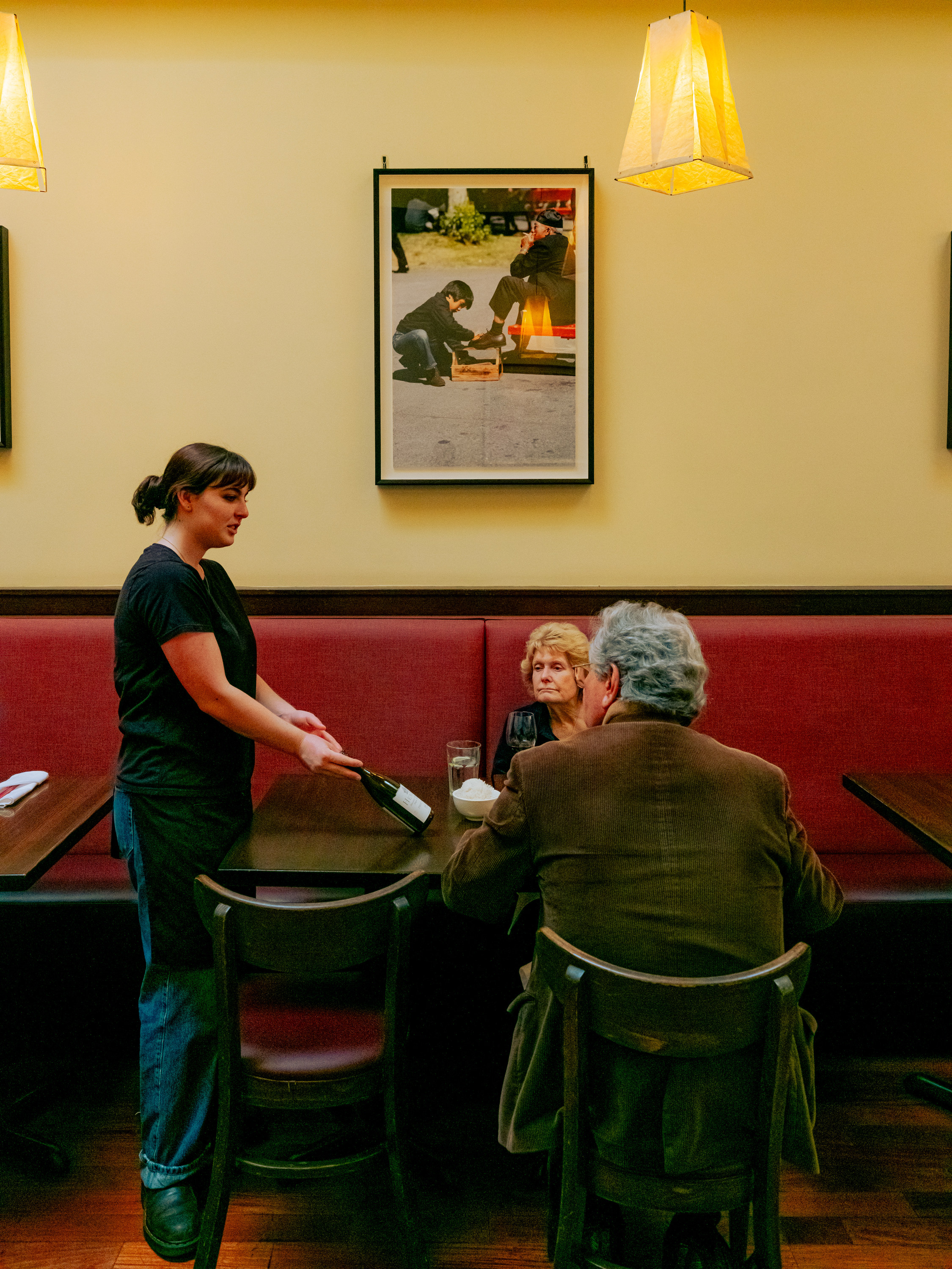 A waitress presents a wine bottle to a seated couple at a restaurant. A framed photo and warm light fixtures hang on a wall behind them.