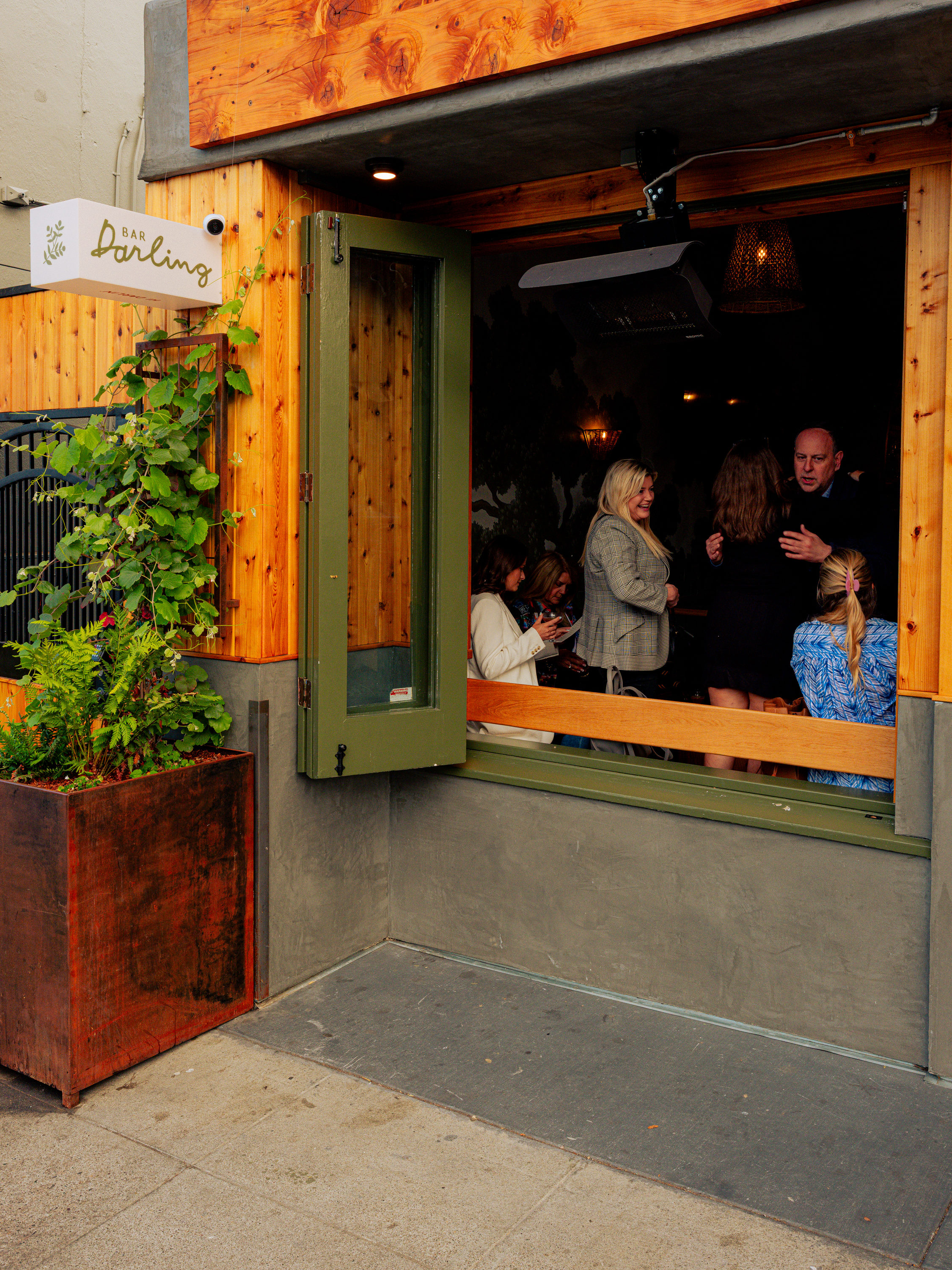 A bar entrance with open green doors, wood accents, and a small plant box. Inside, people are socializing. A sign reads &quot;Bar Darling.&quot;