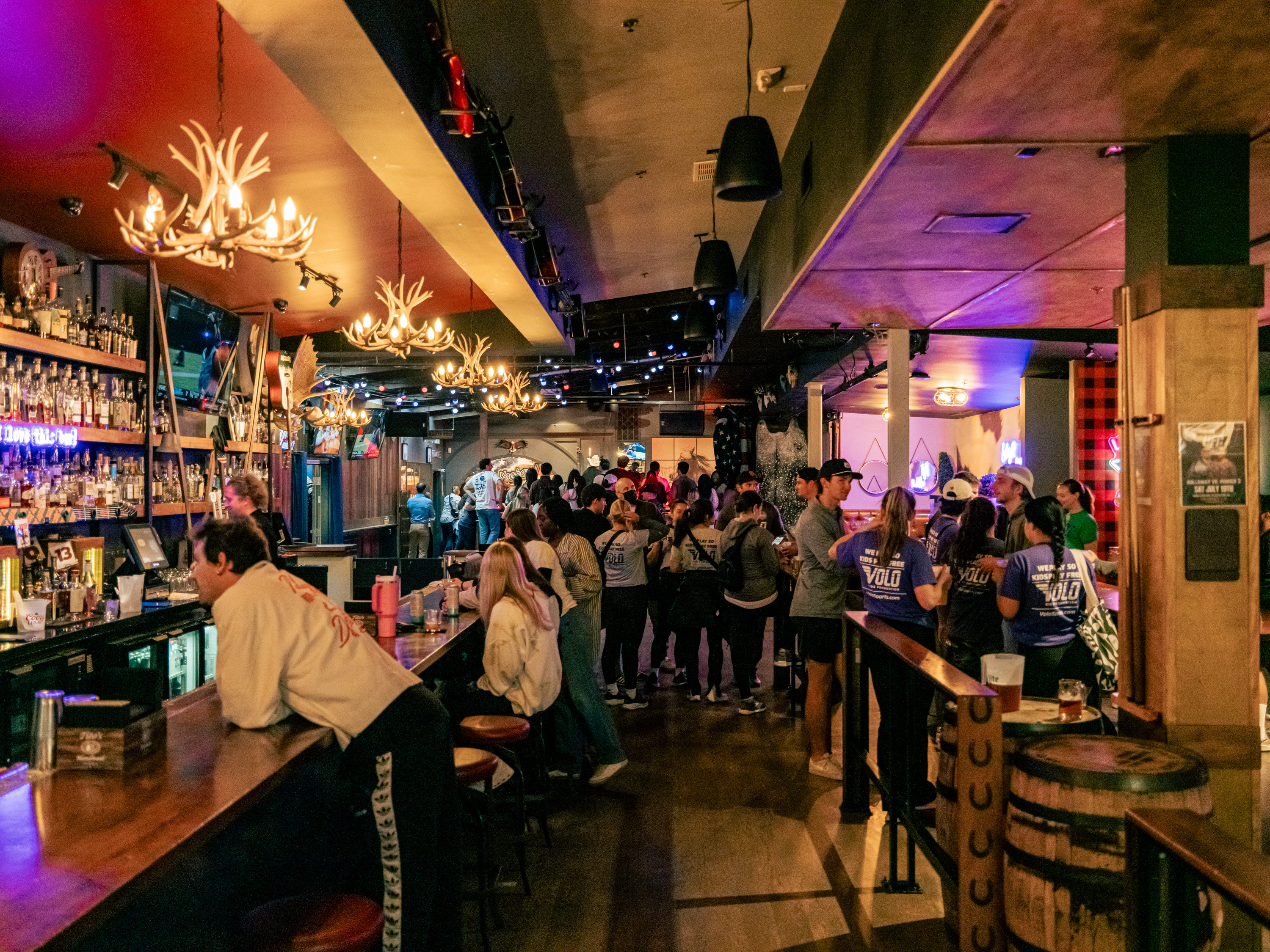 A bustling bar with antler chandeliers, people gathered around, and a fully stocked counter. Patrons chat and socialize amid warm lighting and lively atmosphere.