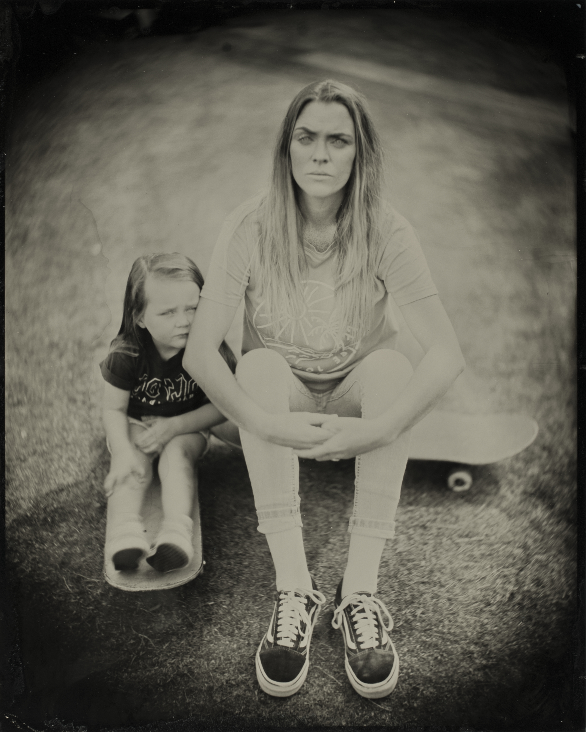 A woman sits on a skateboard, hands clasped, wearing casual clothes and sneakers. A child sits close beside her, also on the skateboard, both serious-faced.