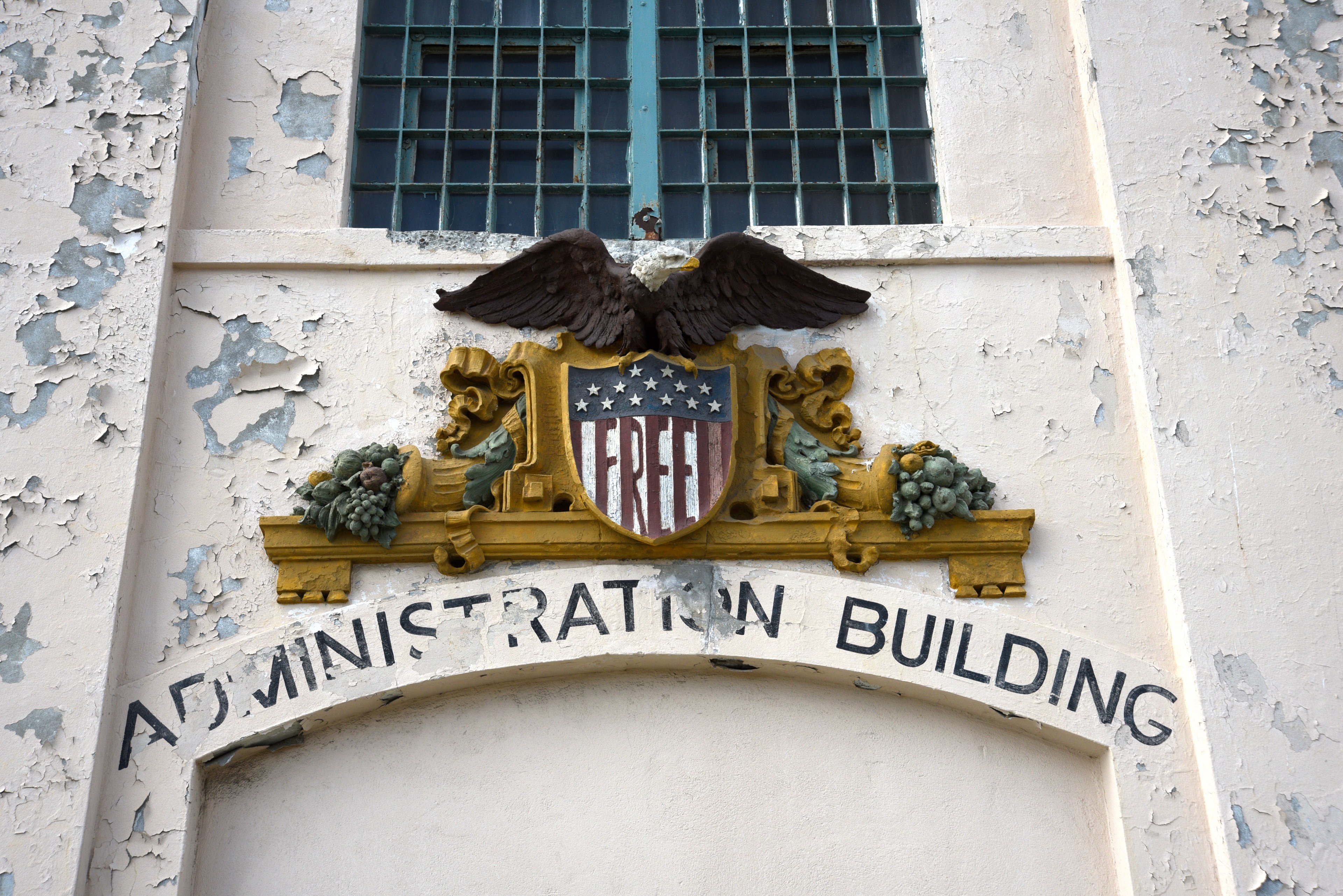 An old building facade features a peeling paint surface, an eagle atop a shield with &quot;FREE,&quot; and an arch labeled &quot;ADMINISTRATION BUILDING&quot; in bold text.